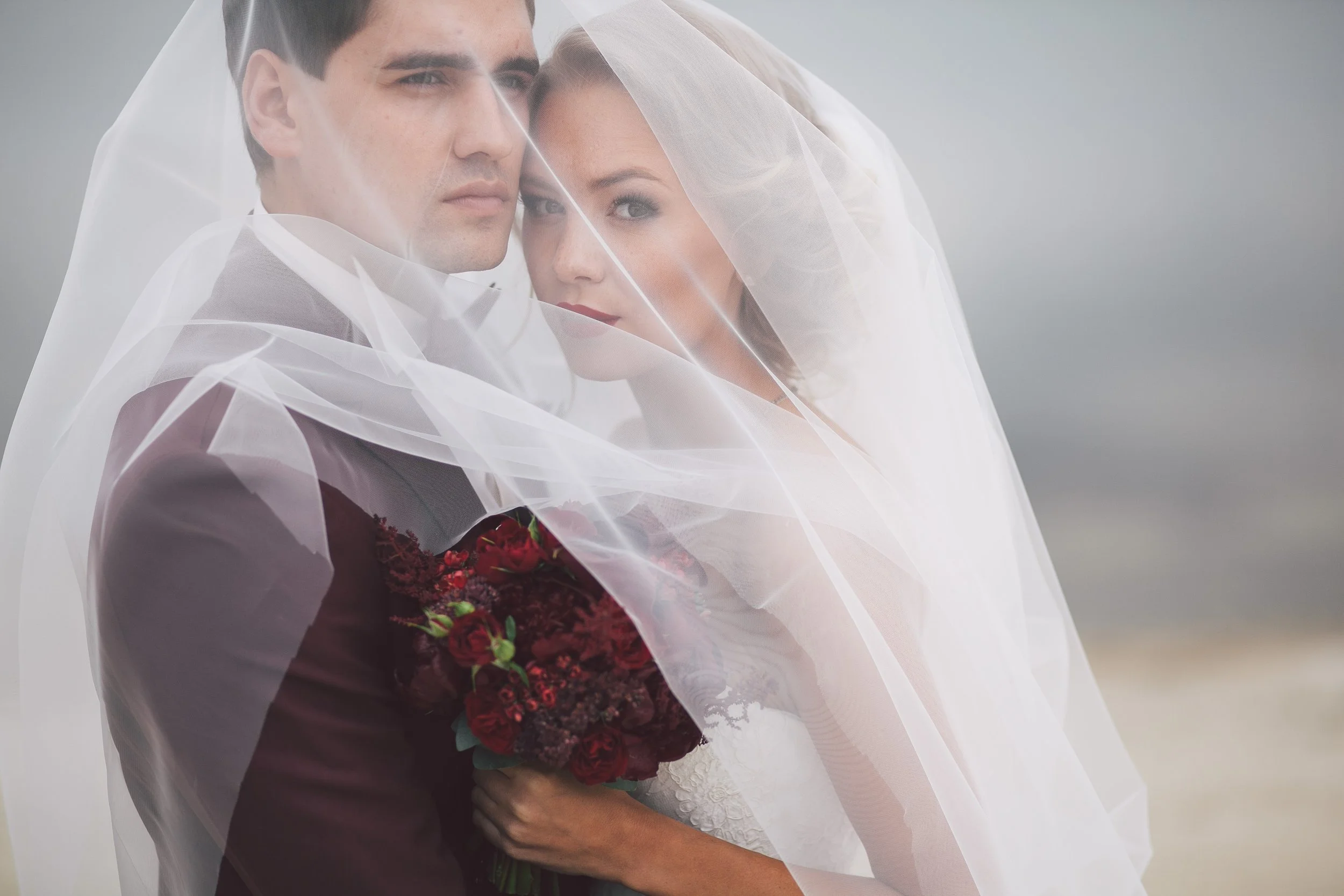 A bride and groom posing together, the bride holding a bouquet of red flowers, both covered with a sheer veil, against a gray background.