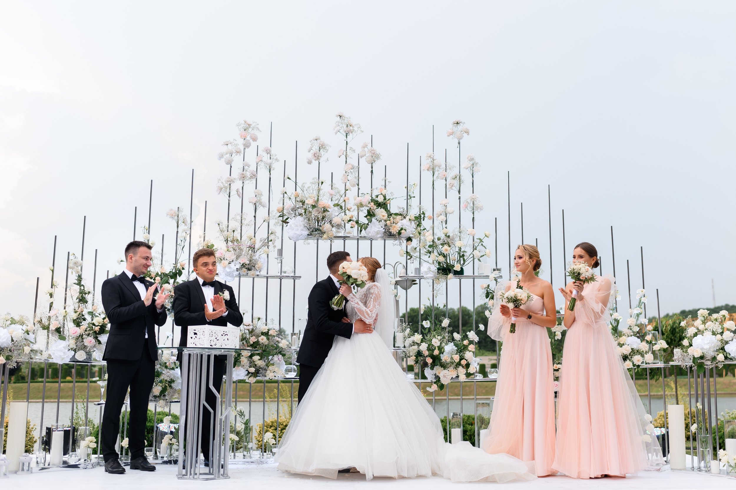 A wedding ceremony with a bride and groom kissing under floral arrangements. Bridesmaids in pink dresses holding bouquets, groomsmen clapping, outdoors with a decorative backdrop of white and pink flowers, and a cloudy sky.