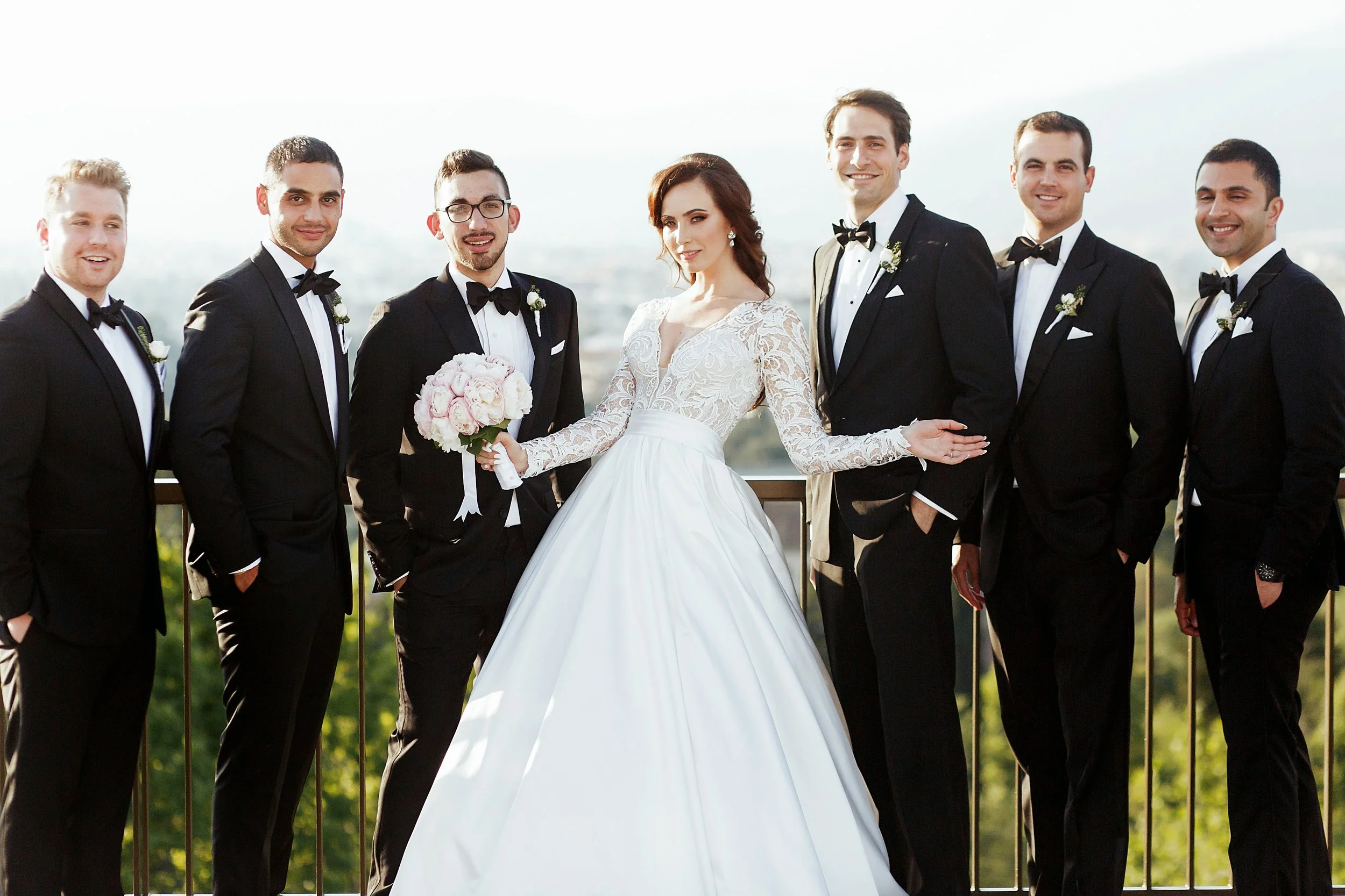 A bride in a white lace wedding dress holding a bouquet of pink and white flowers, standing with six men in black tuxedos with bow ties, smiling outdoors against a bright sky.