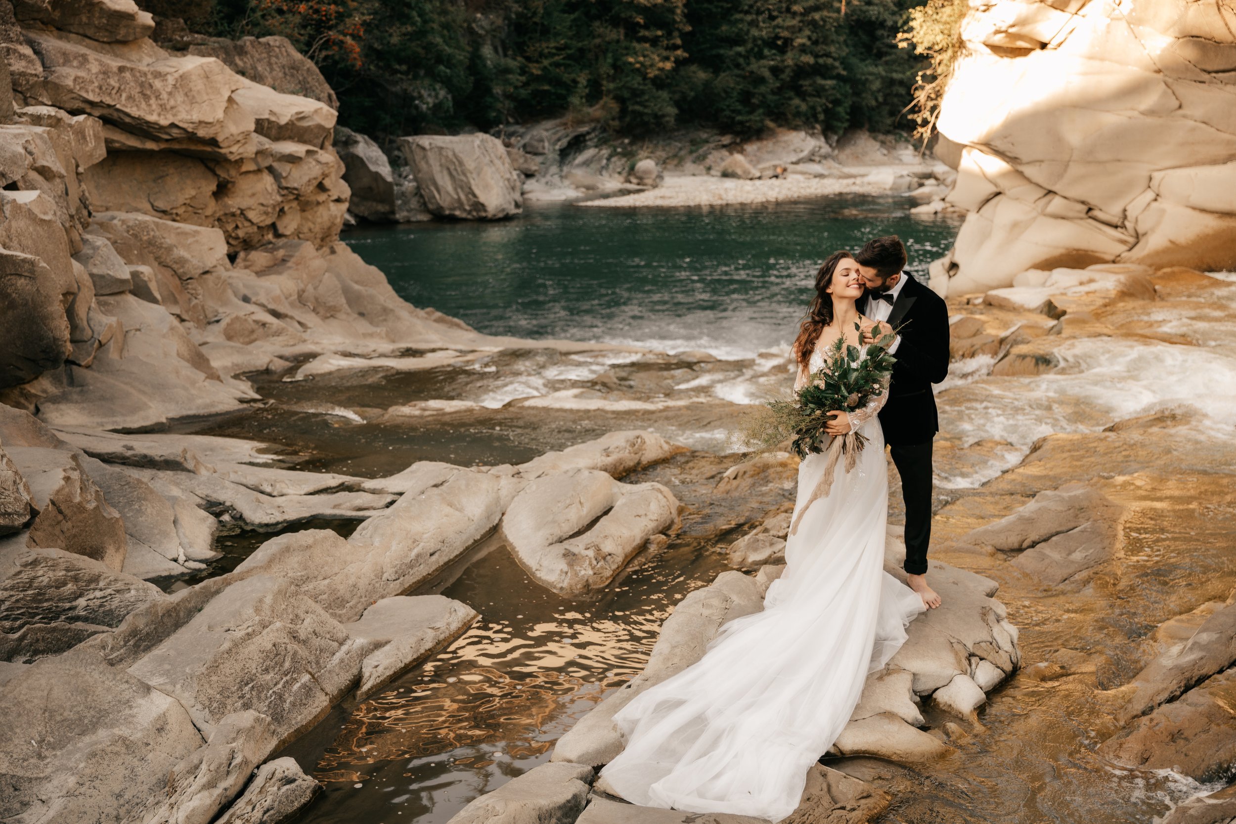 A bride and groom standing together on a rocky riverbank, with the bride holding a bouquet of greenery and the groom kissing her on the cheek. The scene is outdoors near a river with large rocks and a backdrop of forested hills.