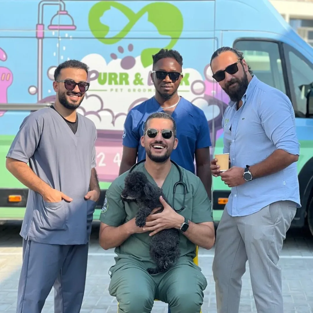 Four men in scrubs and one man in a blue shirt with sunglasses, holding a black puppy, standing in front of a pet food truck that reads 'Purr & Pooch' with colorful illustrations of pets and medical symbols, outdoors on a sunny day.