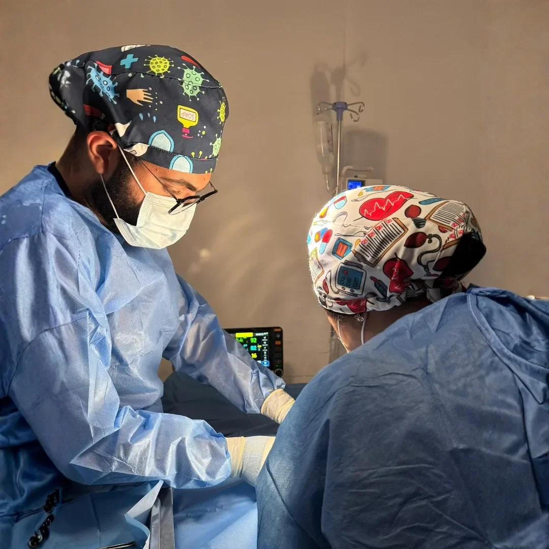 Two medical professionals wearing scrubs, masks, and colorful surgical caps are performing a procedure in a hospital room. A medical monitor is visible in the background.