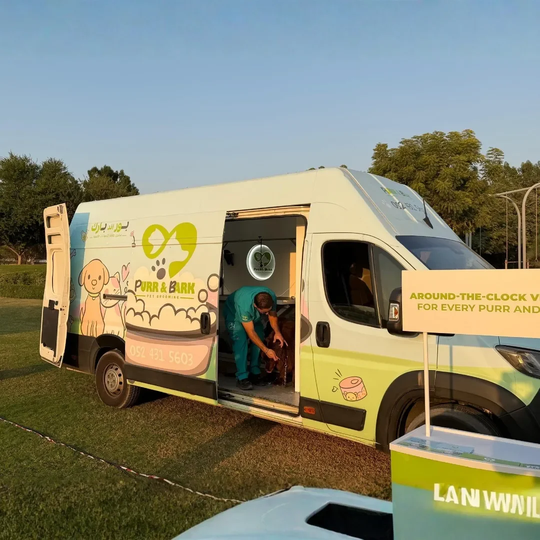 A pet grooming van labeled 'Purr & Bark' parked on a grassy area with a groomer inside grooming a dog, and a sign that reads 'Around-the-Clock Vet for Every Purr and Bark'.