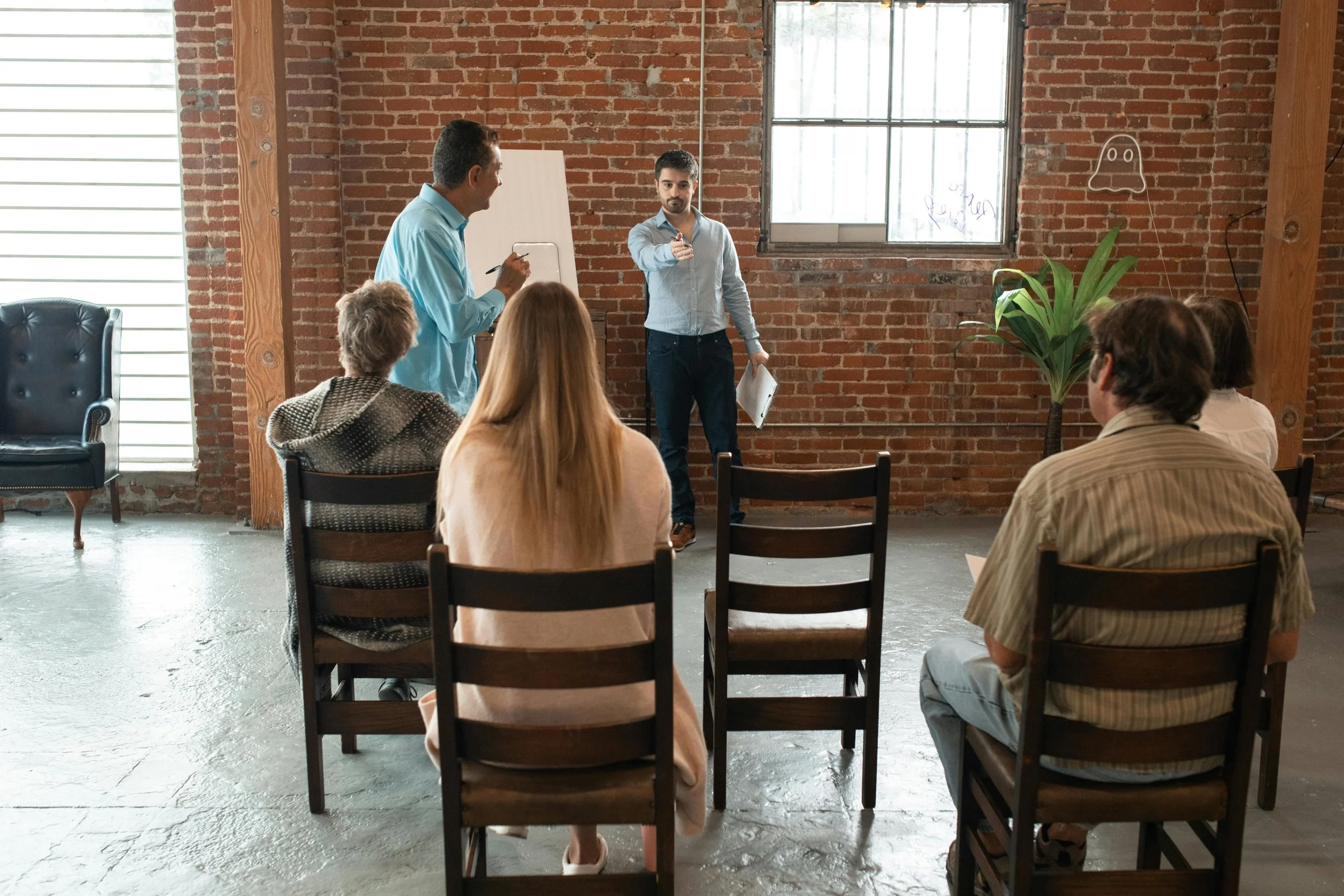 A group of people attending a presentation in a brick-walled room with large windows, seated on wooden chairs, and a man standing at the front pointing at the audience.