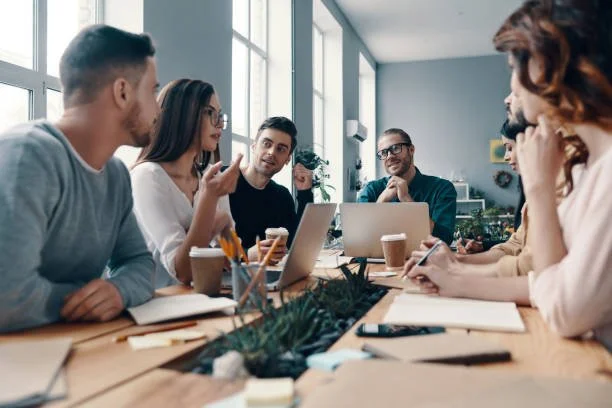 A group of professionals having a meeting in an office, sitting around a table with laptops, notebooks, and coffee cups, engaged in discussion.