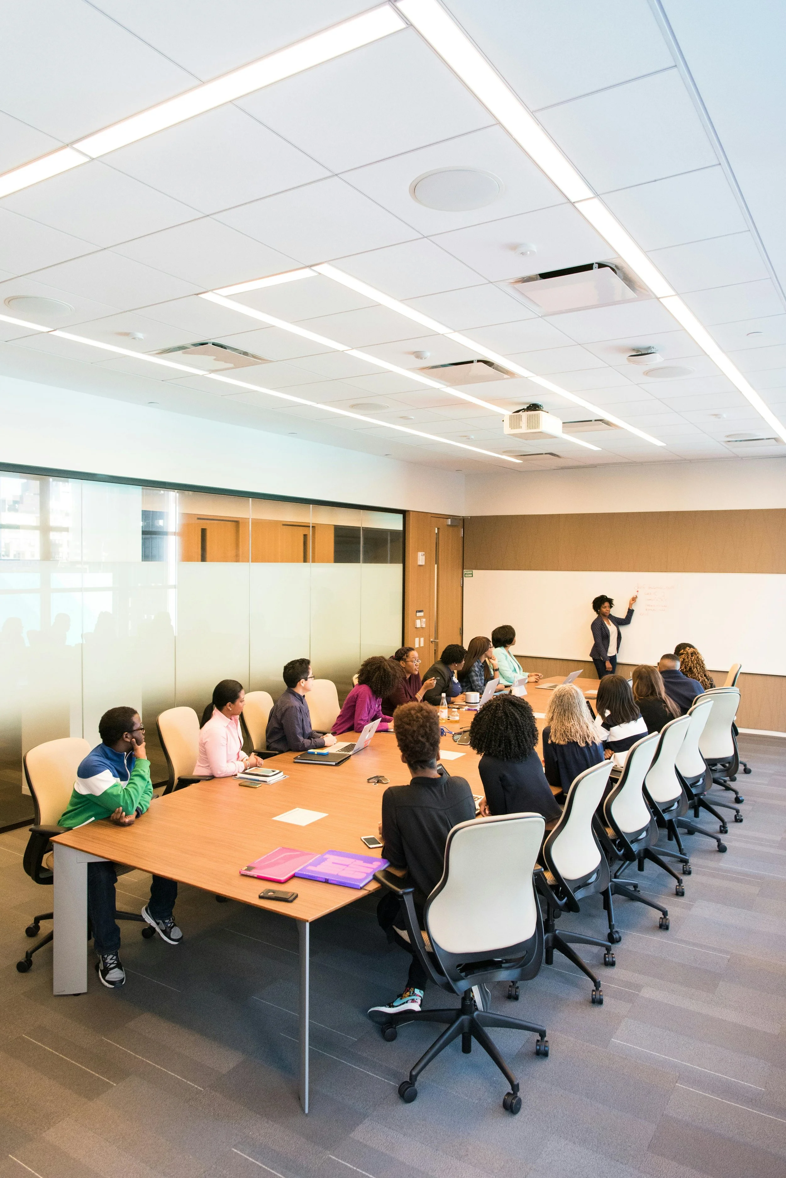 A diverse group of people in a conference room attending a presentation by a woman standing at the whiteboard.