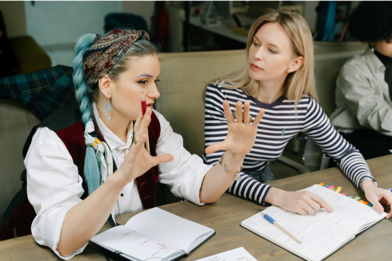 Two women having a discussion at a table with notebooks, one gesturing with her hands and the other listening attentively.