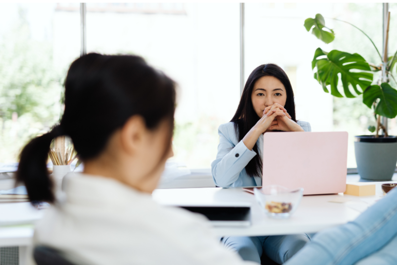 Businesswoman in light blue suit sitting at a desk with a pink laptop, talking with another woman with dark hair in a white top in a bright office with large windows and potted plant.