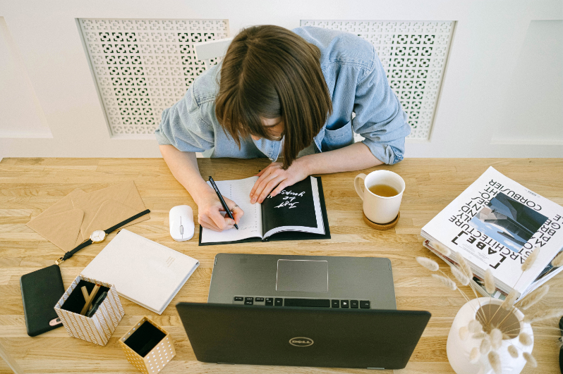 A woman sitting at a desk writing in a notebook with a pen, surrounded by a laptop, a coffee mug, a book, and various office supplies.