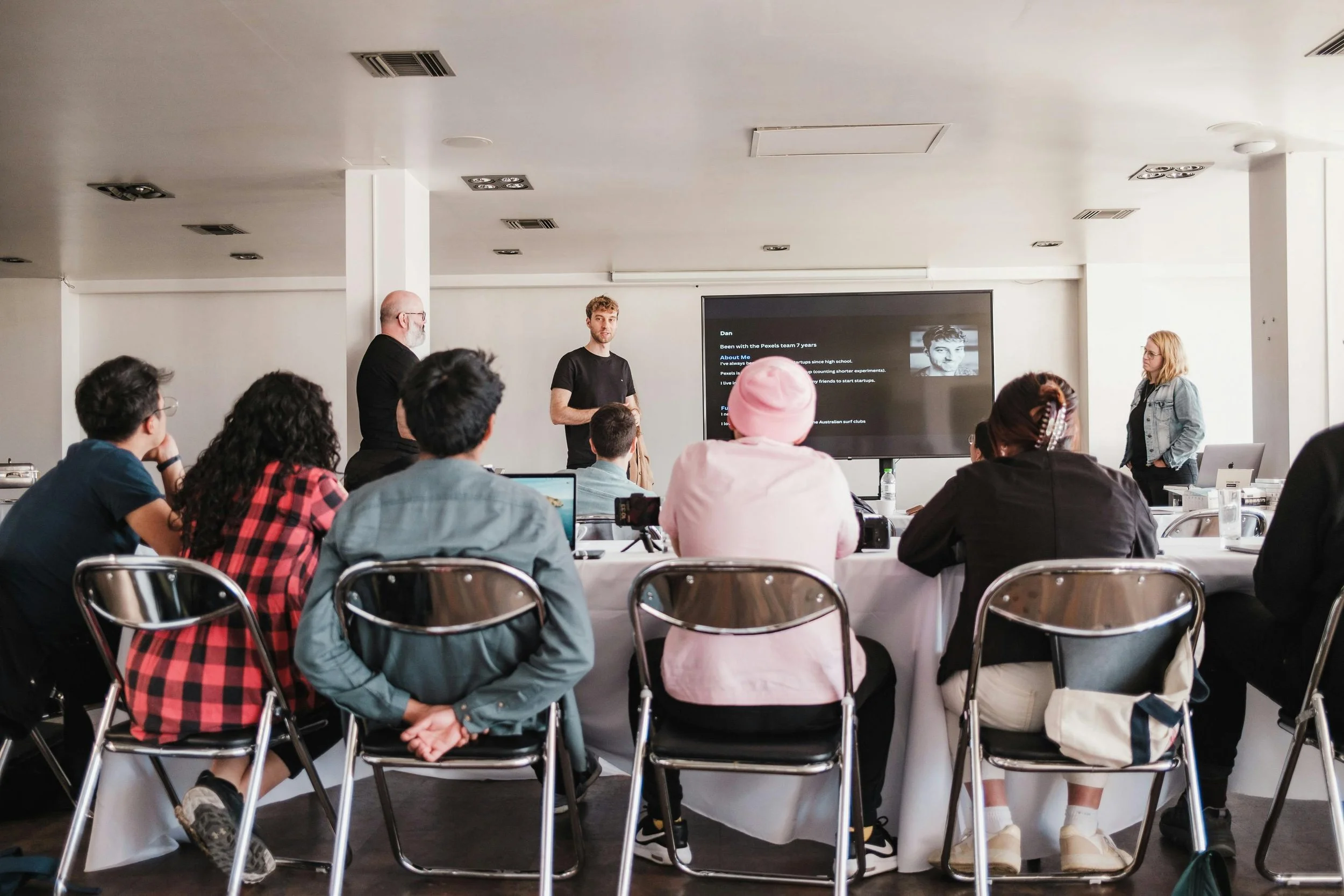 People attending a presentation or workshop in a conference room, sitting around a U-shaped table, focusing on two presenters at the front; one male and one female, with a screen displaying content behind them.