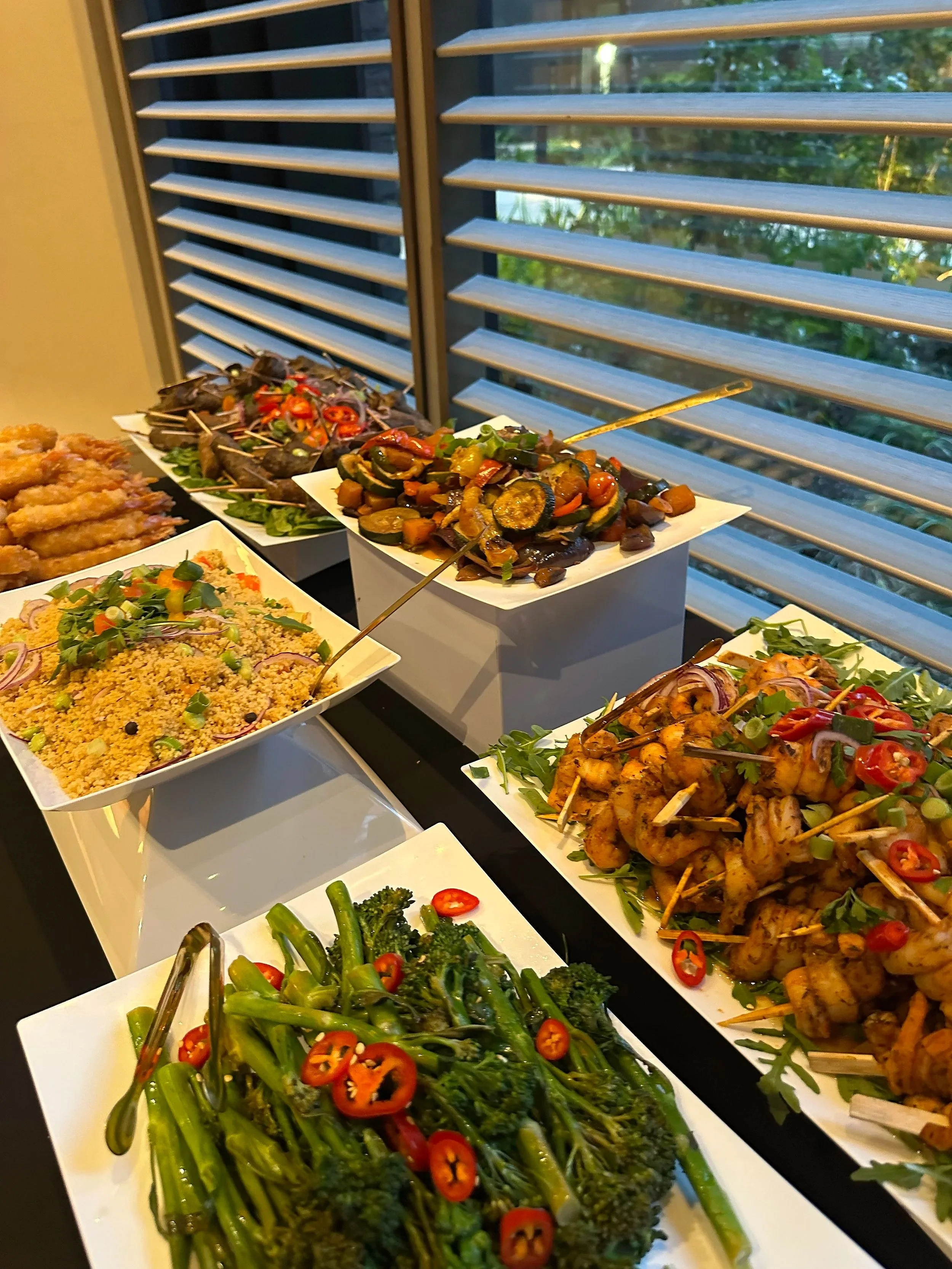 Assorted grilled vegetables including broccoli and red peppers, and skewered chicken, served at a buffet table near a window with blinds.