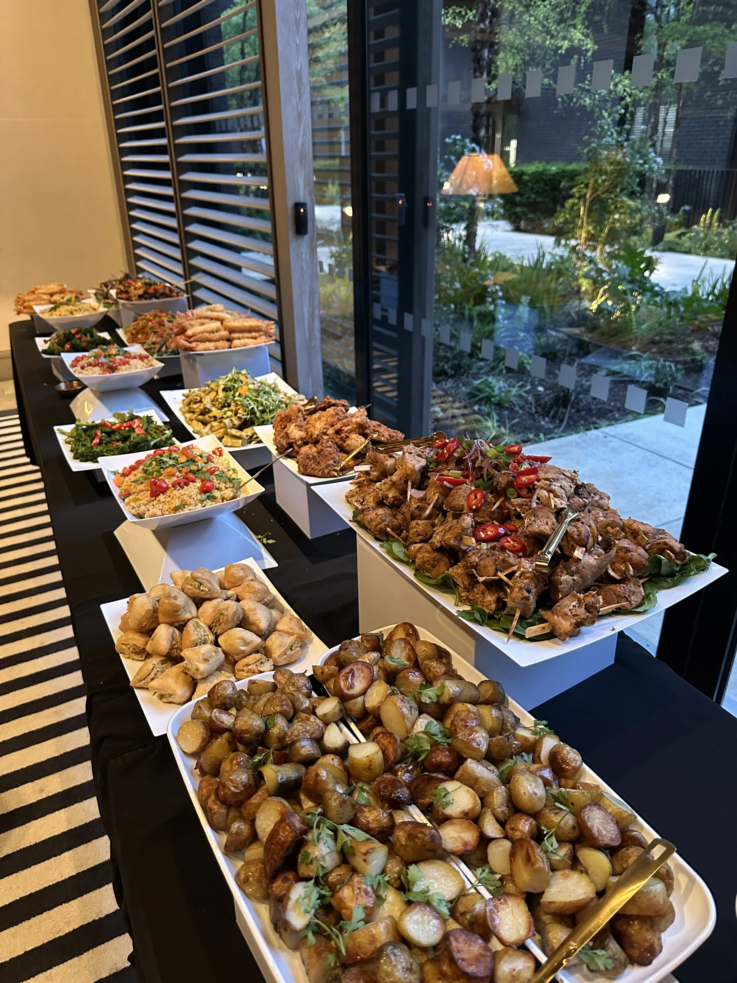 A buffet table with various appetizers including skewered meats, stuffed potatoes, salads, and fried foods, set near a window with a garden view.