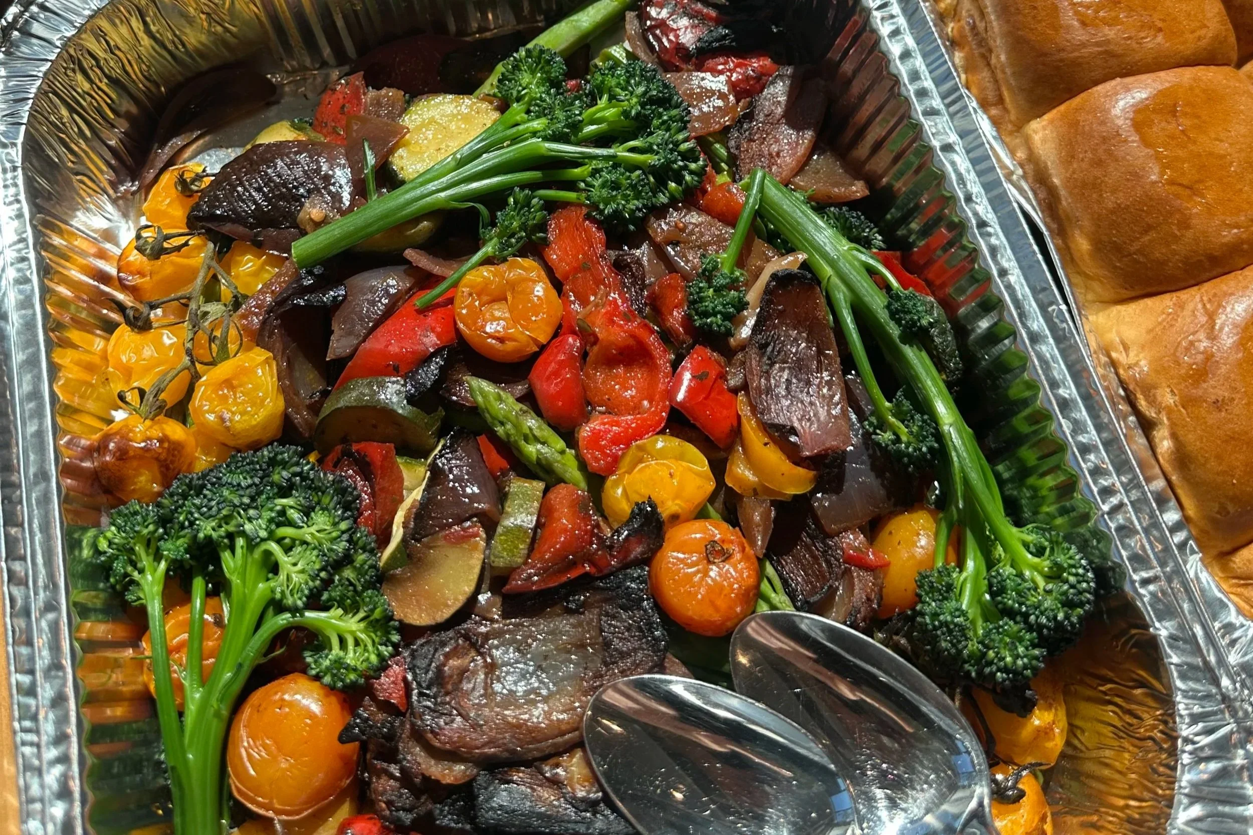 Assorted roasted vegetables including cherry tomatoes, broccoli, red and green peppers, zucchini, and carrots in a foil tray with mashed potatoes and bread rolls on the side.
