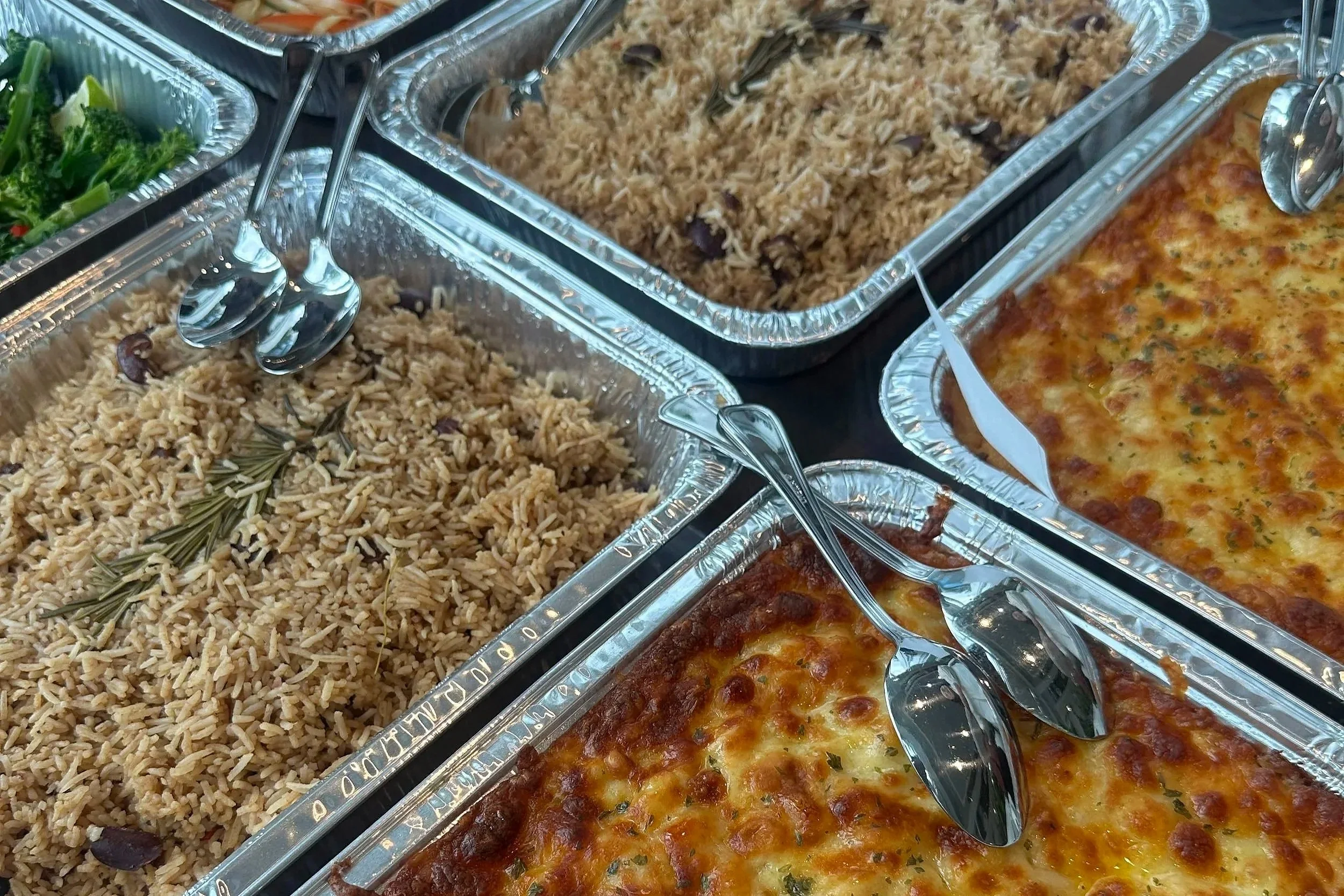 Trays of food in a buffet style setting including rice dishes, green salad, and baked casserole, all in aluminum containers with serving utensils.