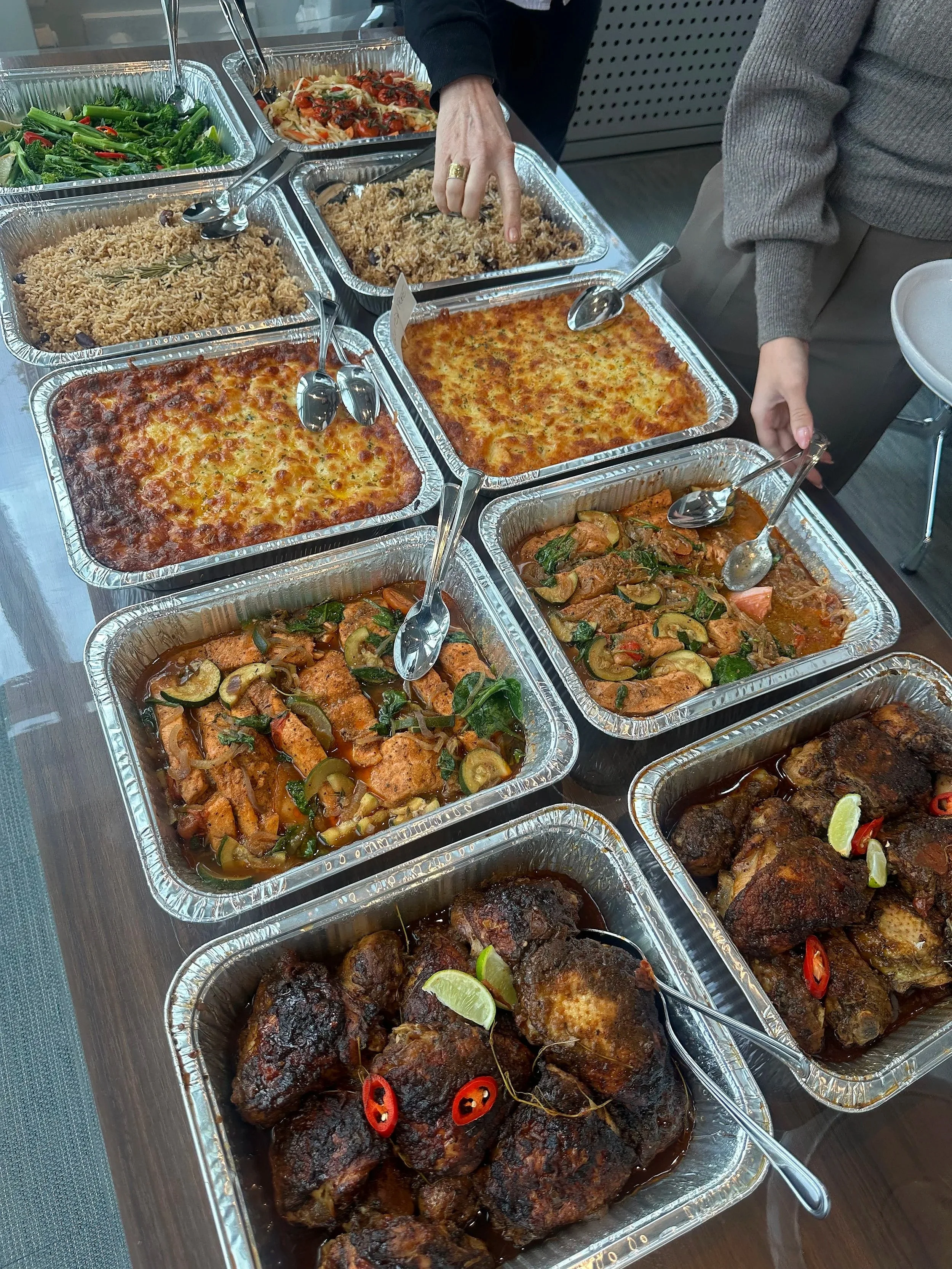 A buffet table with several aluminum trays filled with various cooked dishes, including baked chicken, rice, pasta, and vegetable dishes, with a person pointing at one of the trays.