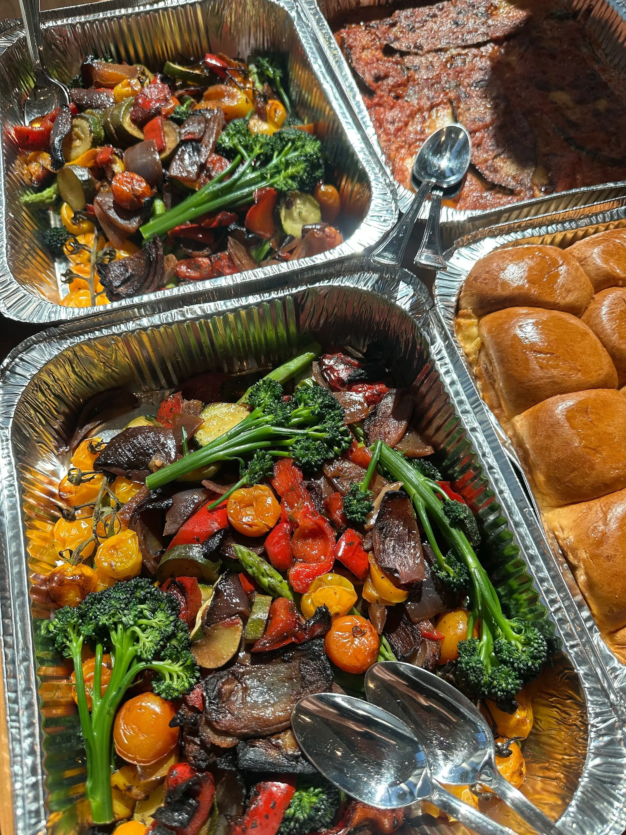 Aluminum foil trays filled with roasted vegetables including cherry tomatoes, broccoli, red and yellow bell peppers, zucchini, and eggplant, with serving spoons.