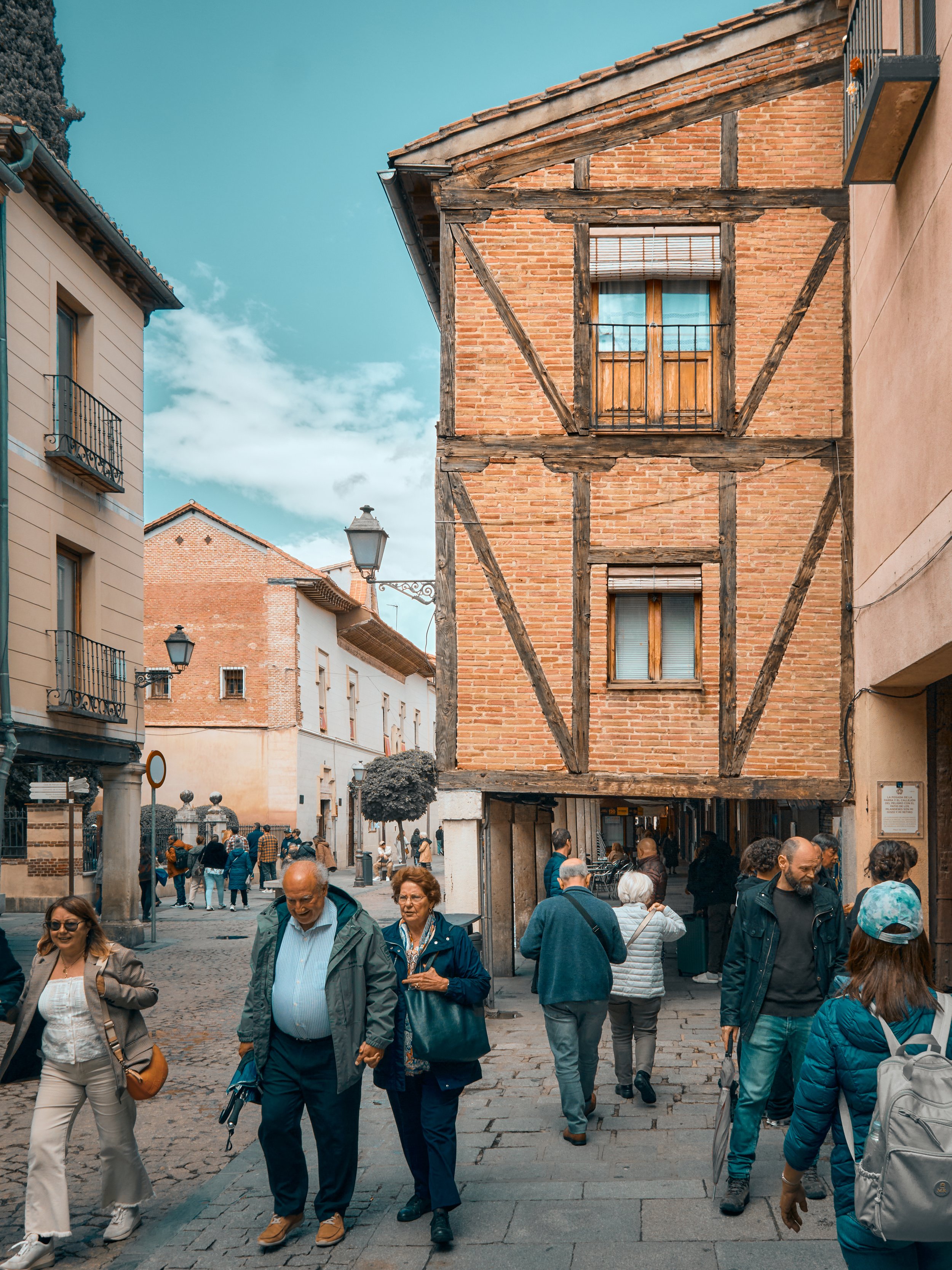 A la calle con personas caminando, edificio de dos pisos de ladrillo y madera en el centro, con casas de color claro y farolas en las calles.