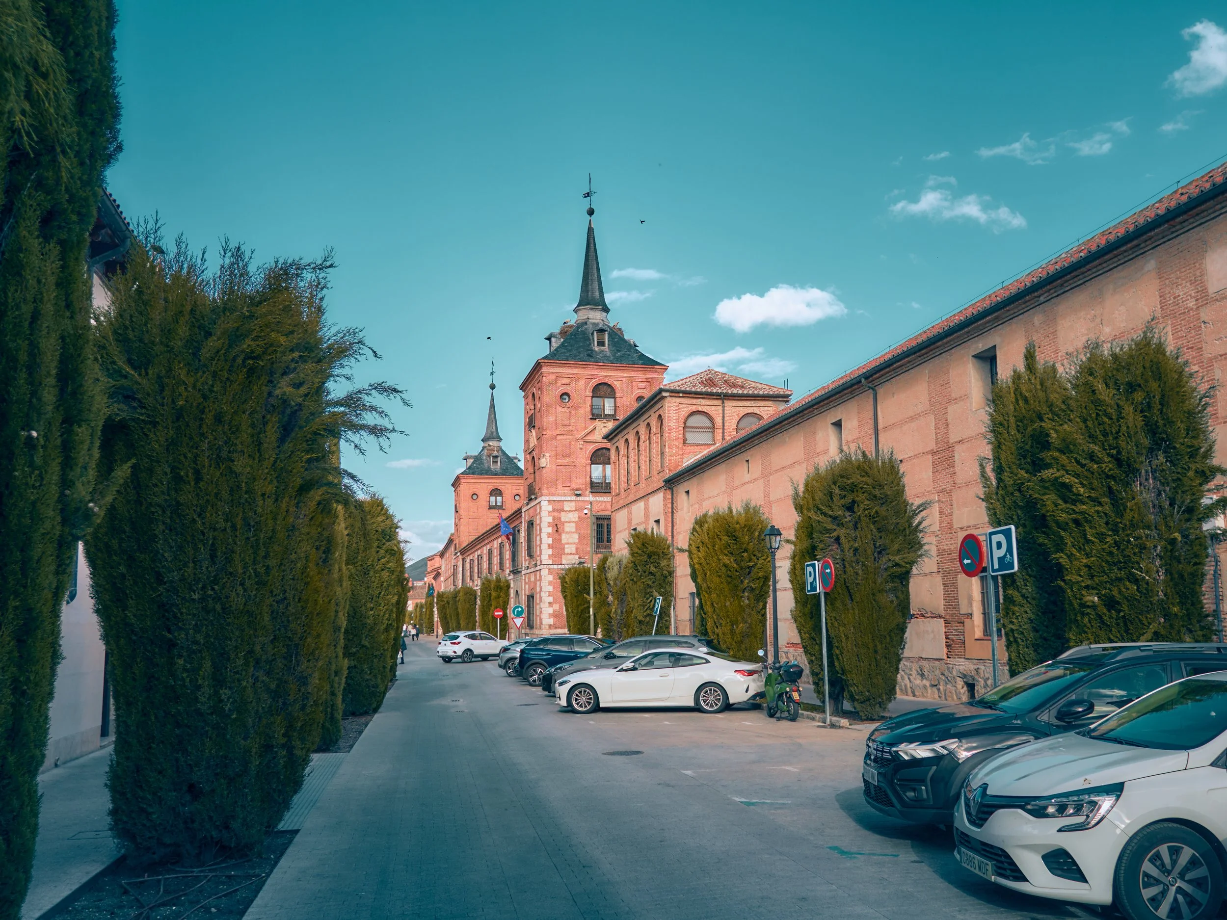 Calzada con árboles y coches estacionados en frente de un edificio histórico con torres y cúpulas, cielo despejado con algunas nubes.