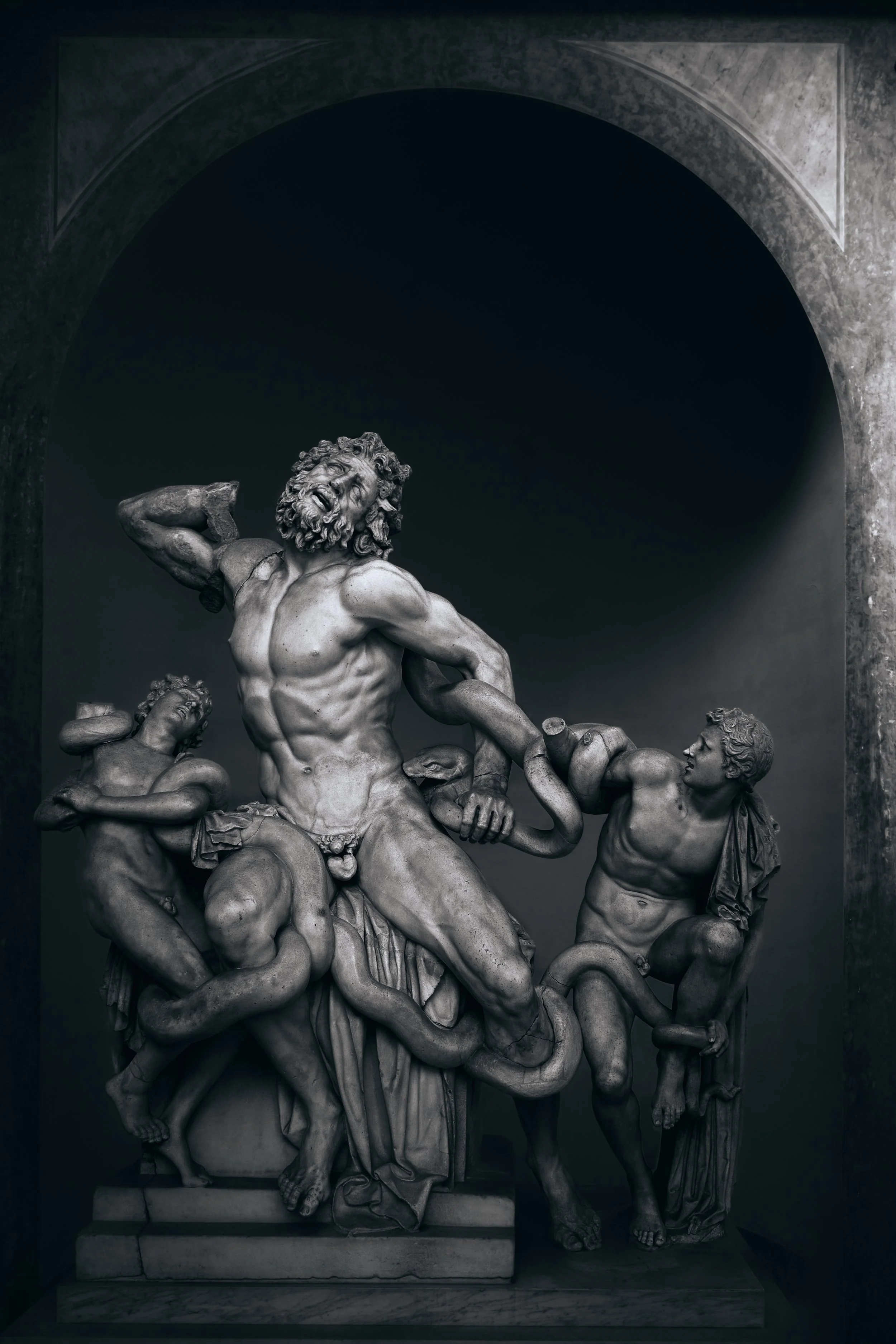 Escultura de mármol en colores grises de un dios griego en medio de una lucha con serpientes y figuras humanas, en un estilo clásico.