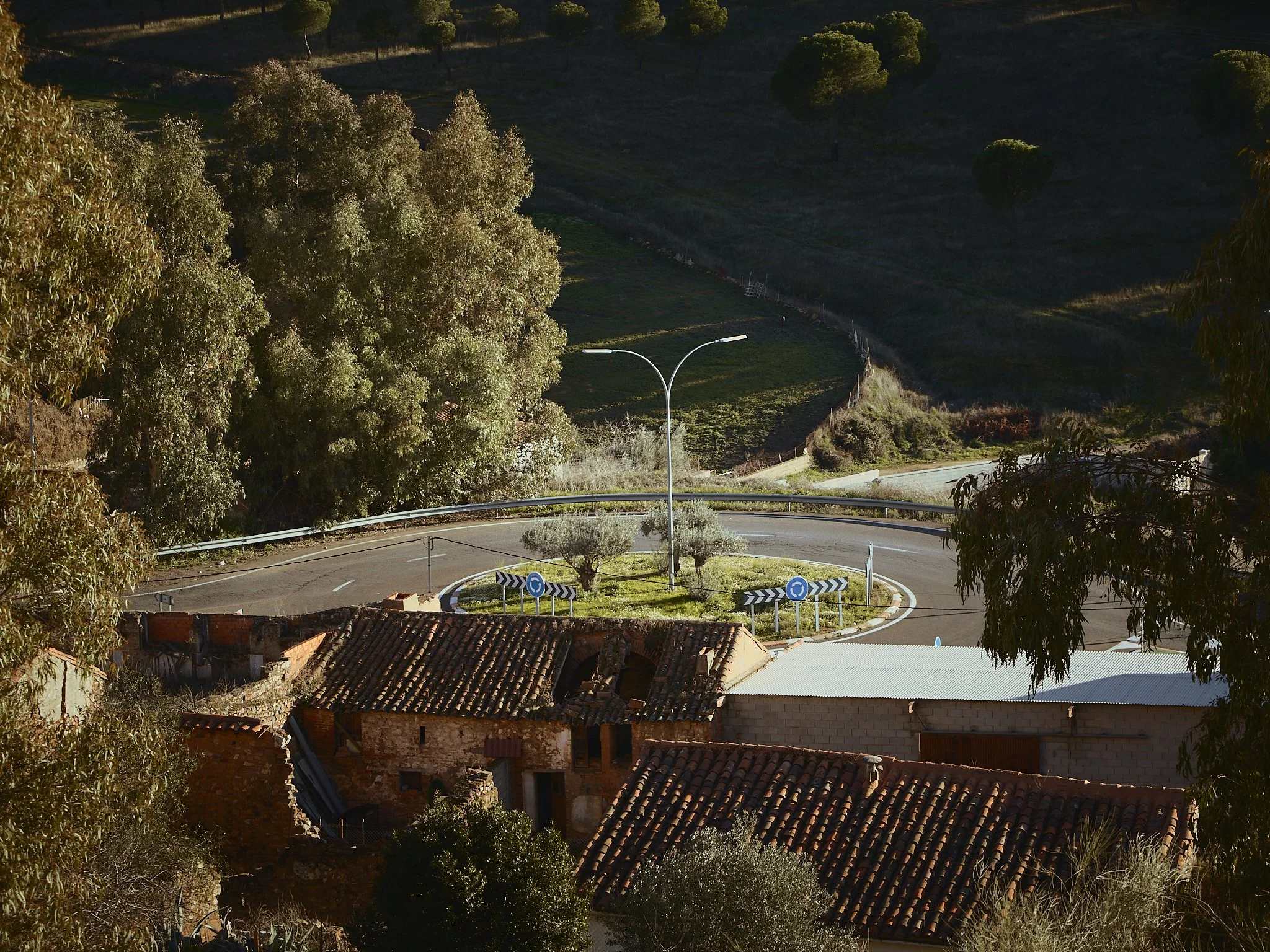 Vista de una rotonda en una zona rural rodeada de árboles y casas con techos de tejas, en un paisaje montañoso con vegetación.