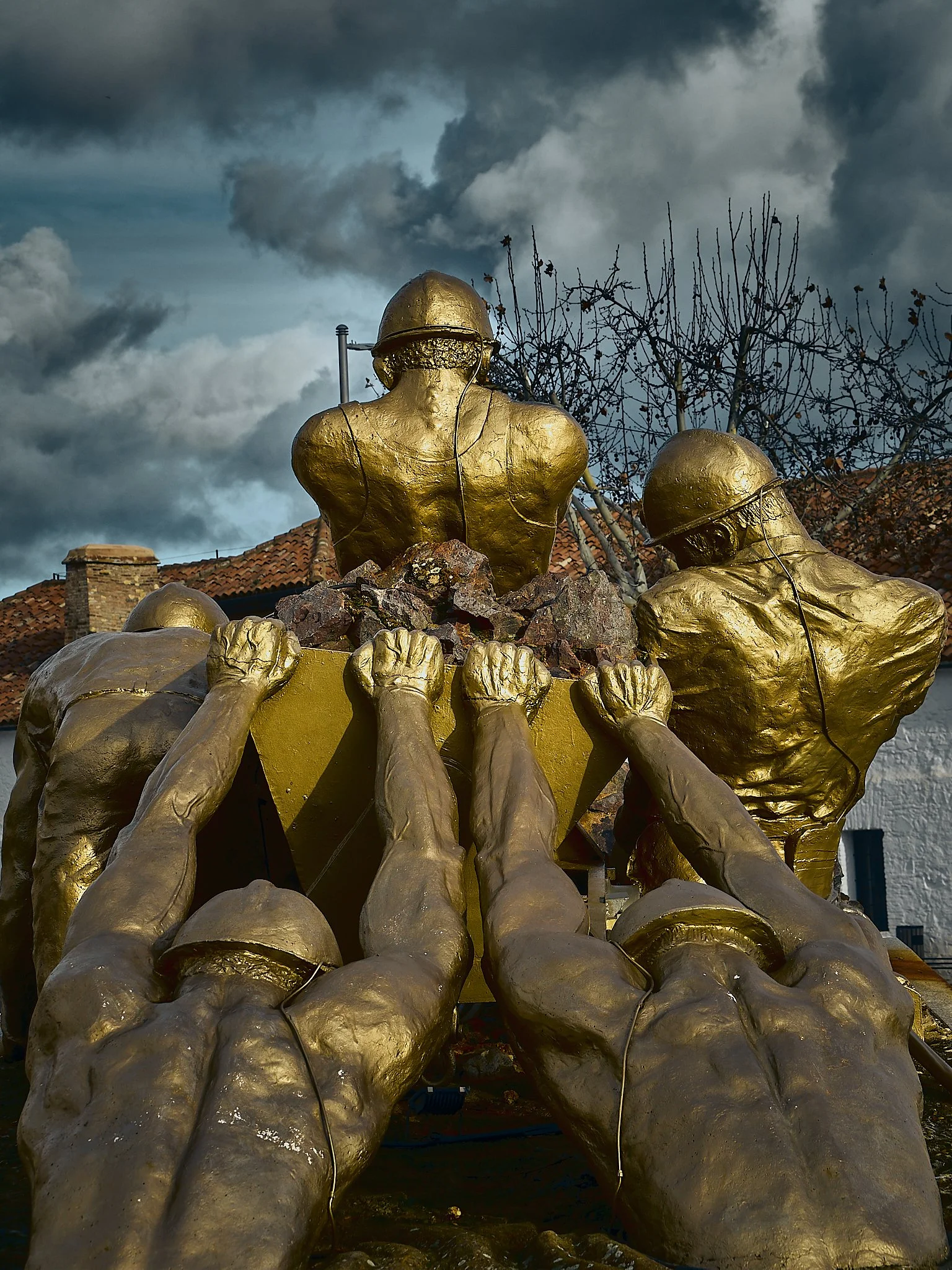 Escultura con figuras de soldados con cascos y manos extendidas pintadas en dorado, con un fondo de cielo nublado y árboles sin hojas.