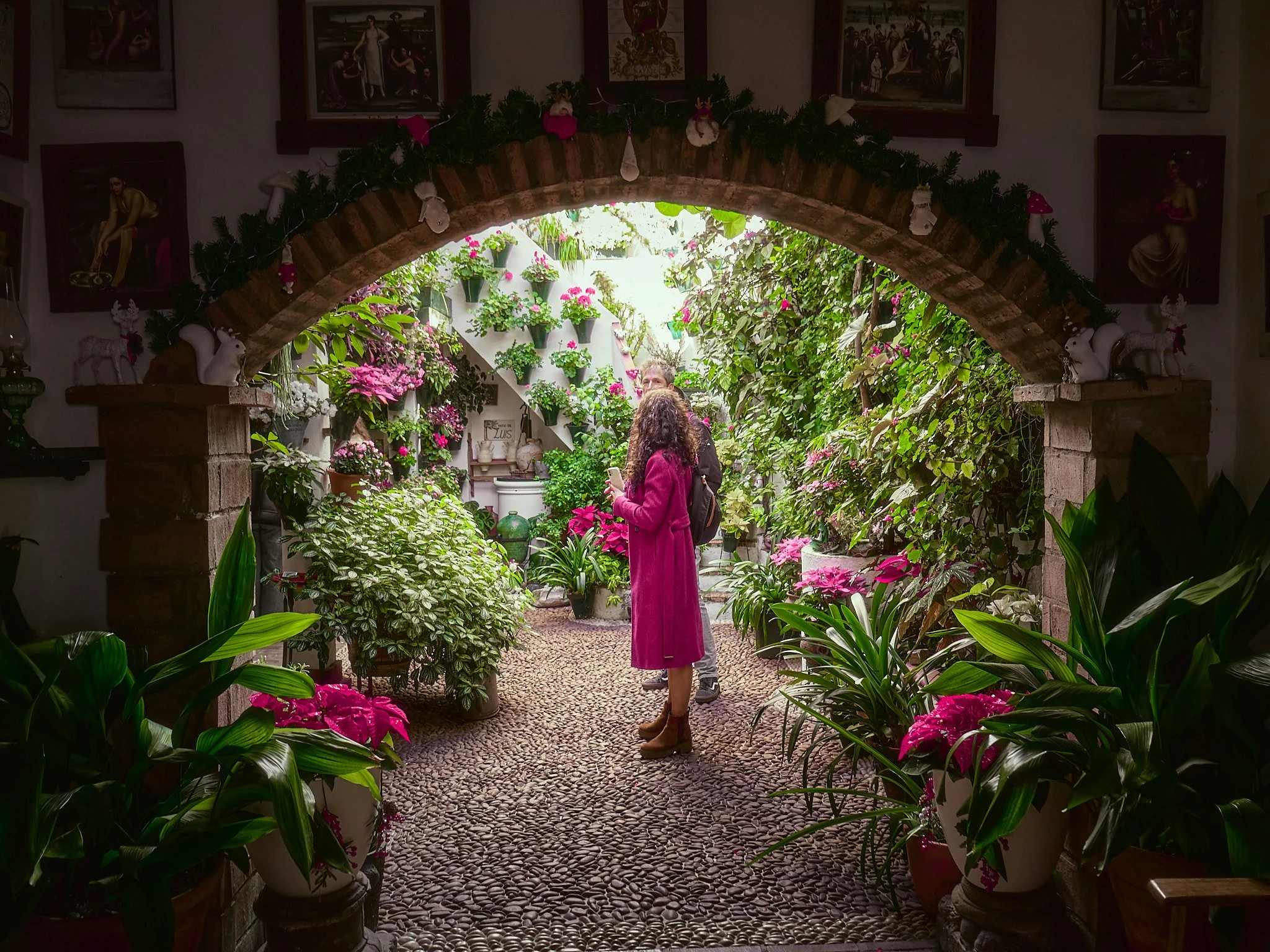 Jardín de interior con muchas plantas y flores, dos personas miran y conversan bajo un arco de ladrillos decorado con adornos navideños. En la parte superior hay cuadros en las paredes.