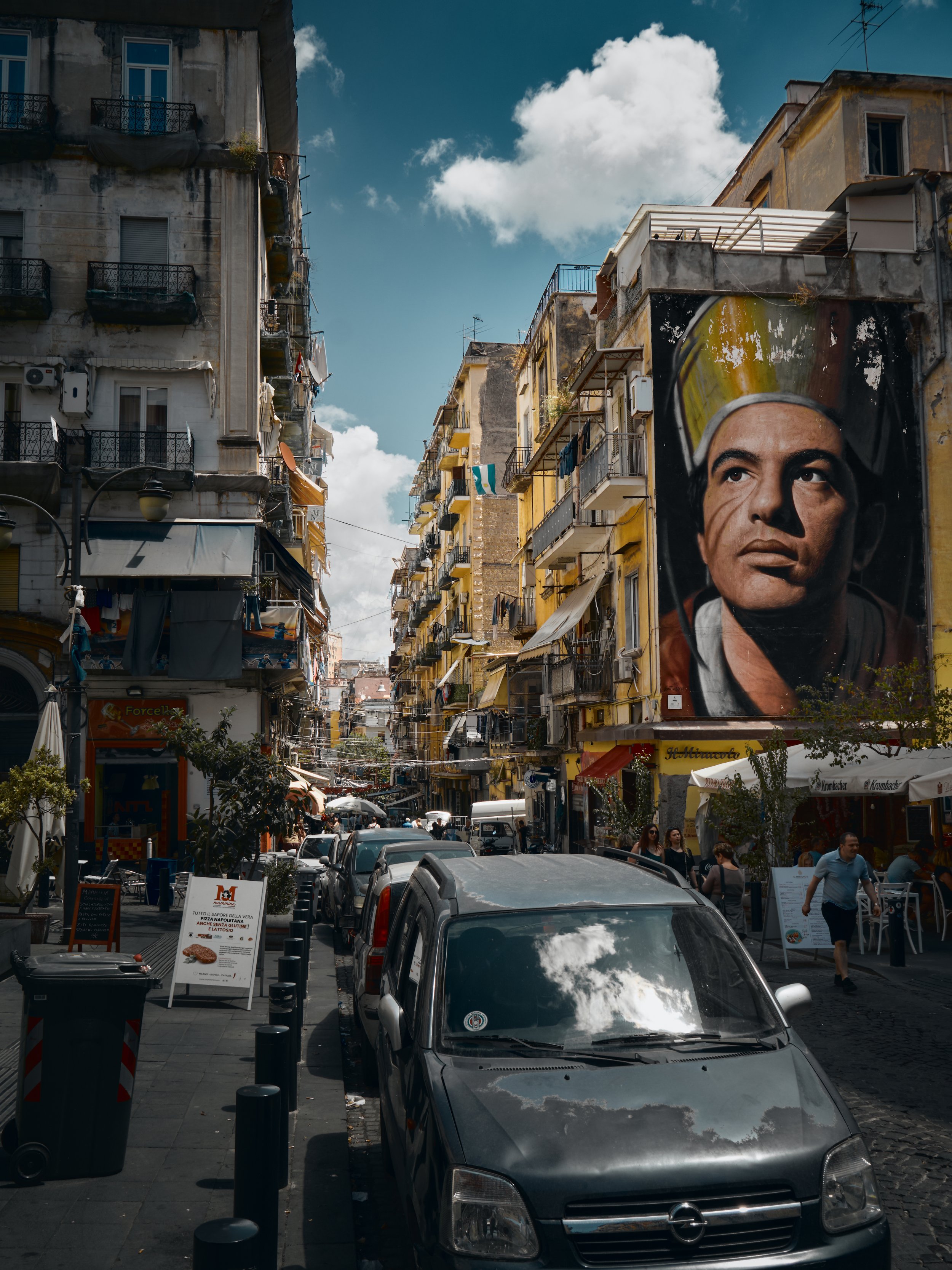 Vista de una calle concurrida en un vecindario urbano con edificios de varios pisos, un gran mural de un hombre con casco y algunos vehículos estacionados.
