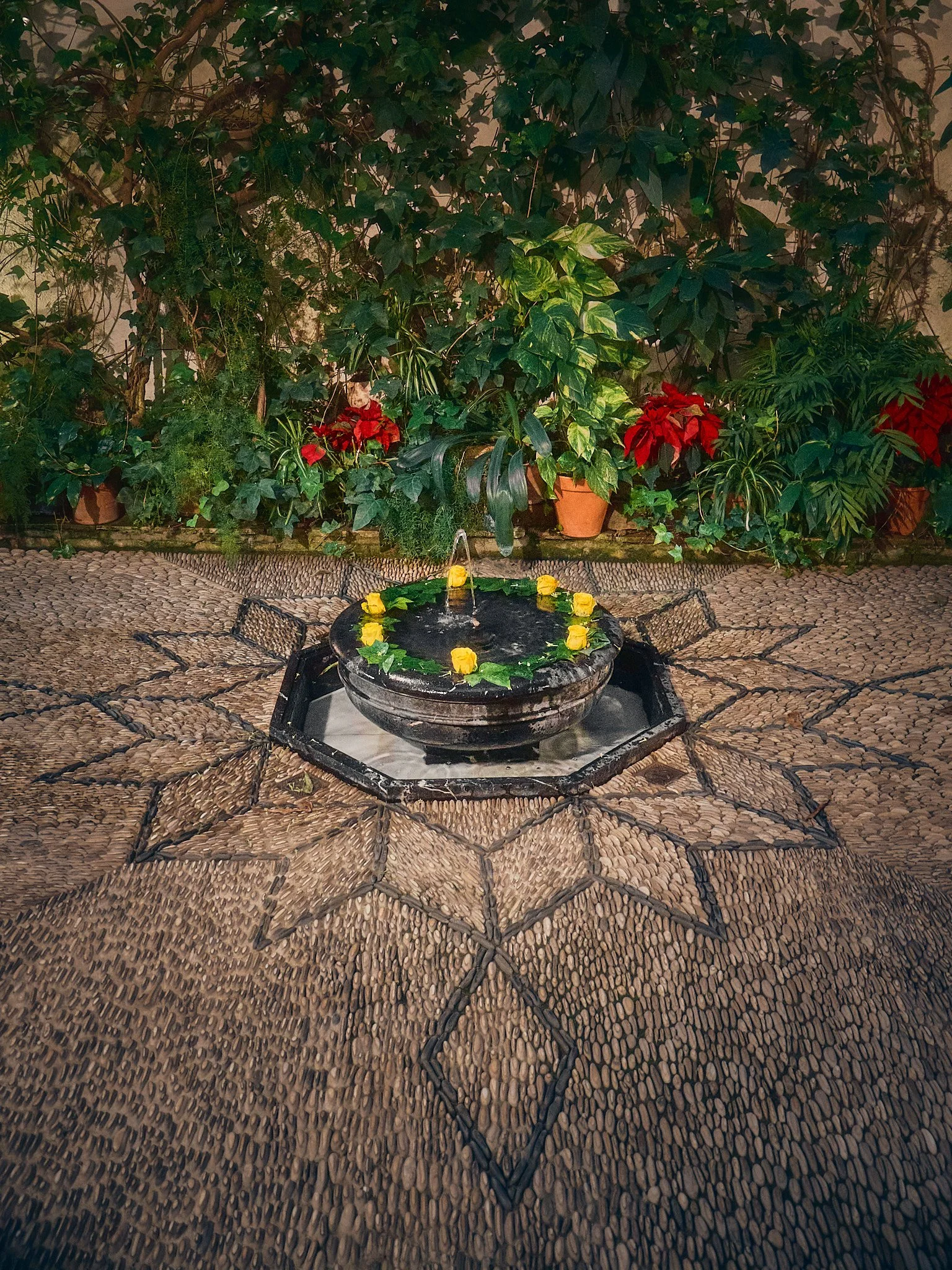 Fuente ornamentada con flores amarillas y hojas verdes en el centro, rodeada de mosaico con patrón en estrella en el piso, y plantas y flores rojas y verdes en el fondo.