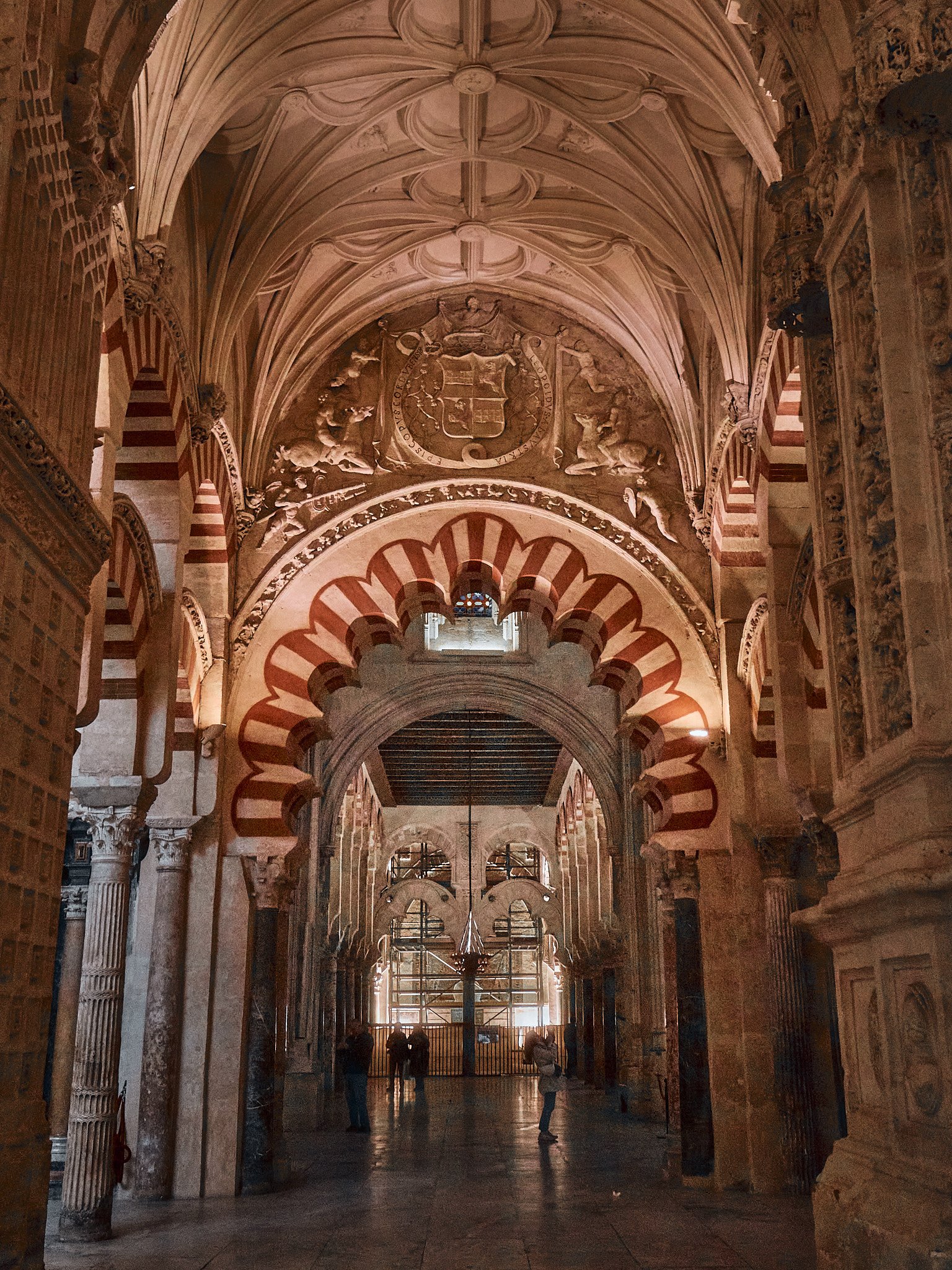 Interior de una iglesia o catedral con techos altos, arcos decorativos rojos y blancos, esculturas y relieves en las paredes, con varios visitantes caminando en el pasillo central.