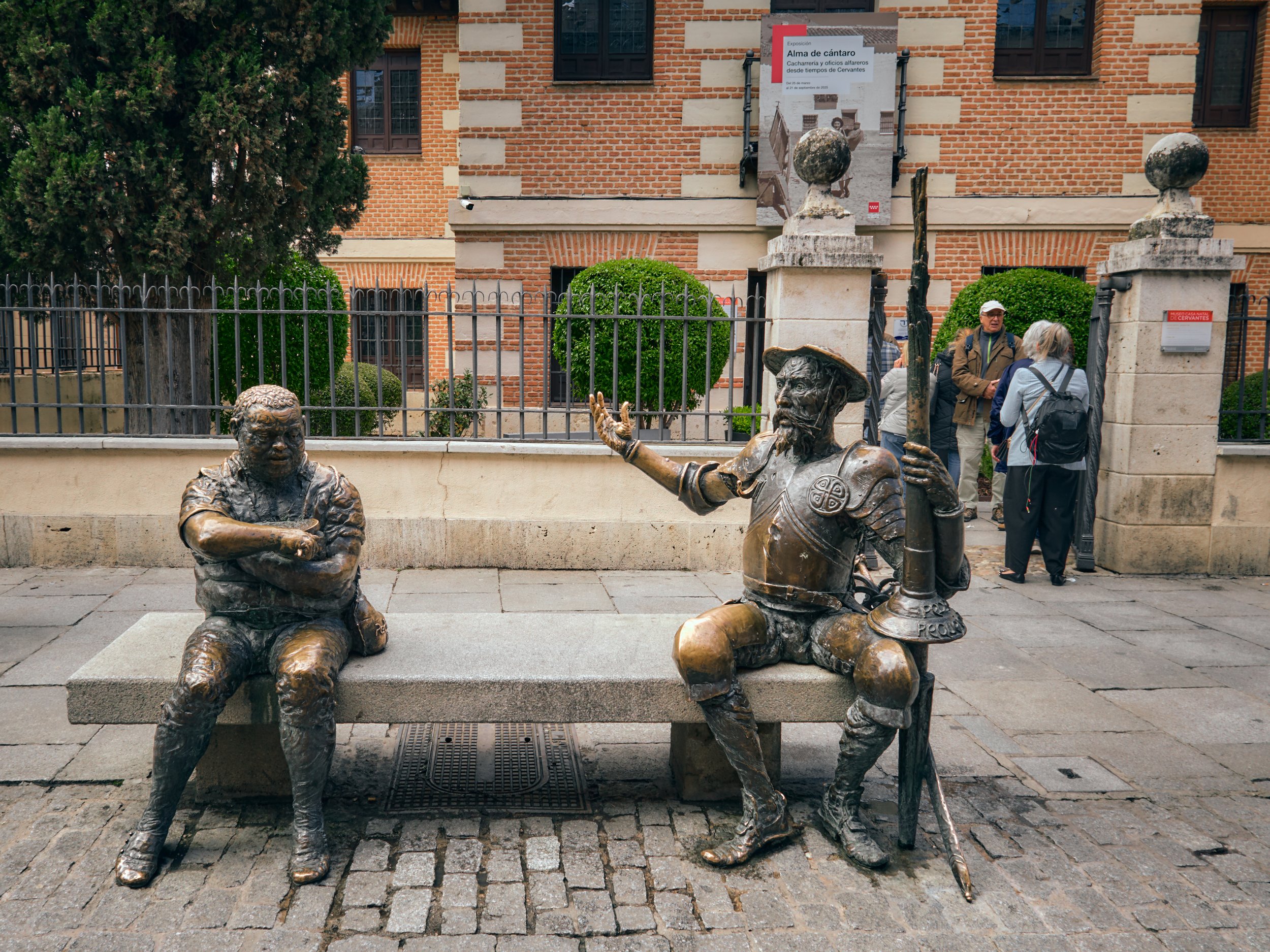 Escultura en la calle de dos personajes históricos sentados en un banco, uno con brazos cruzados y el otro con un sombrero y un arma, en un entorno urbano con personas al fondo.