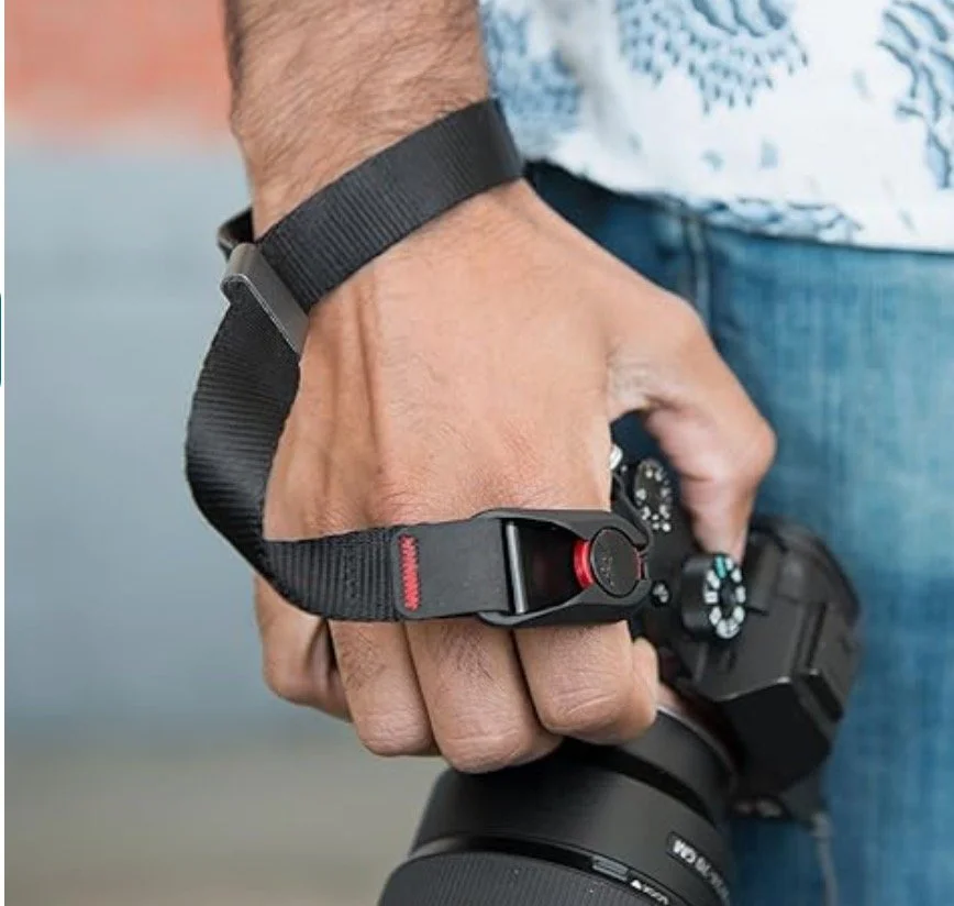 Una mano sosteniendo una cámara y usando una cinta de seguridad con un clip rojo en la muñeca.