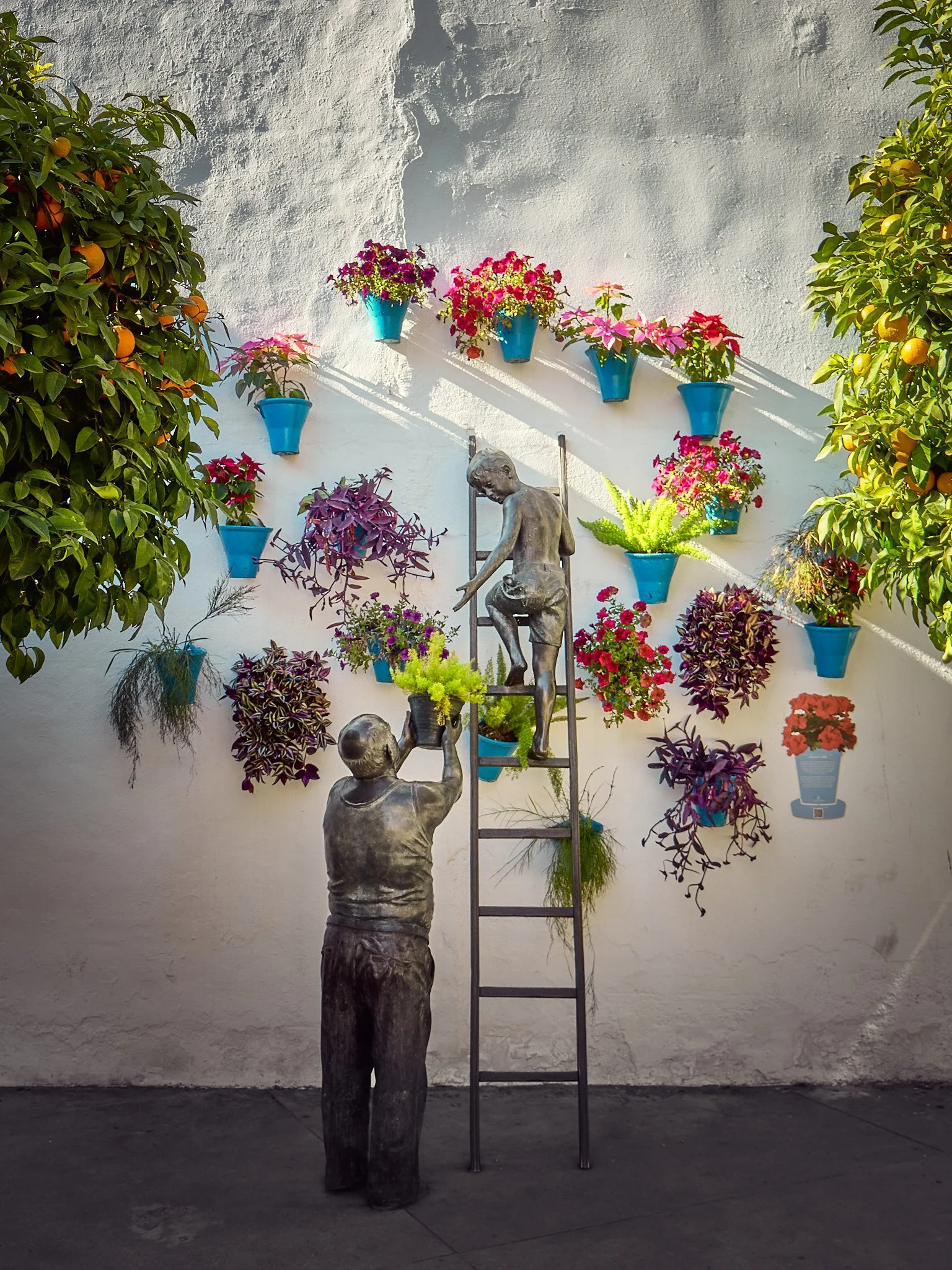 Esculturas de dos personas ayudándose a subir en una escalera frente a una pared decorada con macetas de flores.