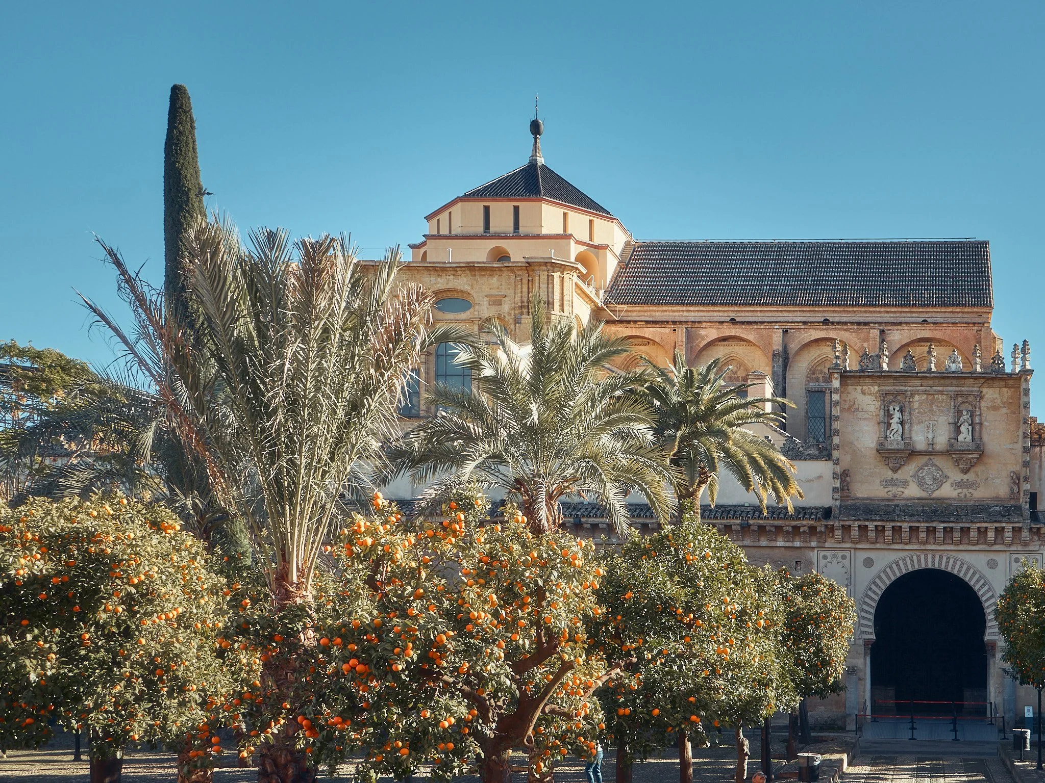 Abadía con árboles frutales y palmeras en primer plano, y edificio histórico con campanario y arquitectura antigua, bajo cielo despejado.