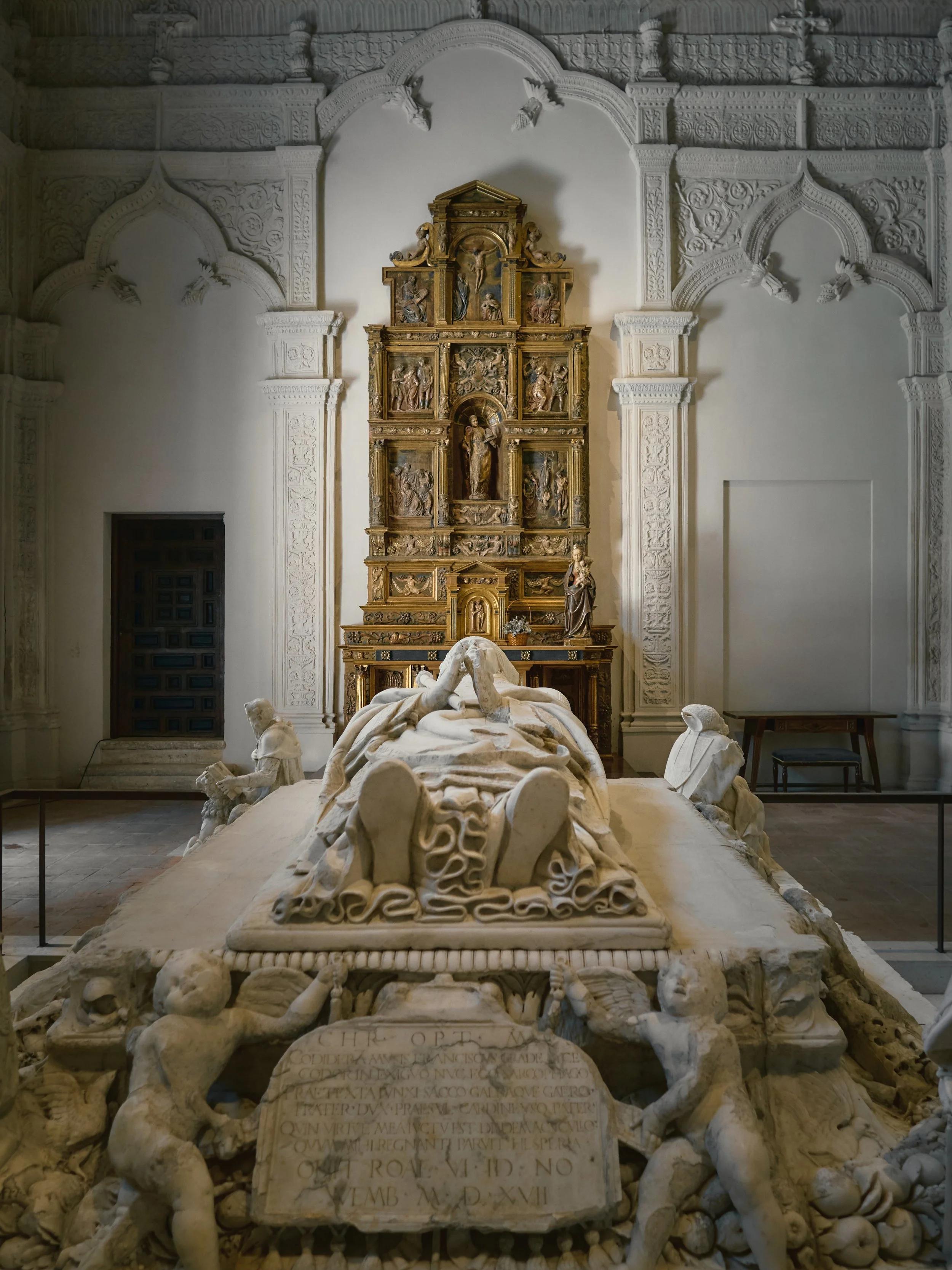 Tumba de mármol con esculturas de niños en la base, en un salón con paredes blancas y detalles arquitectónicos en relieve, y un altar dorado con retablo en el fondo.