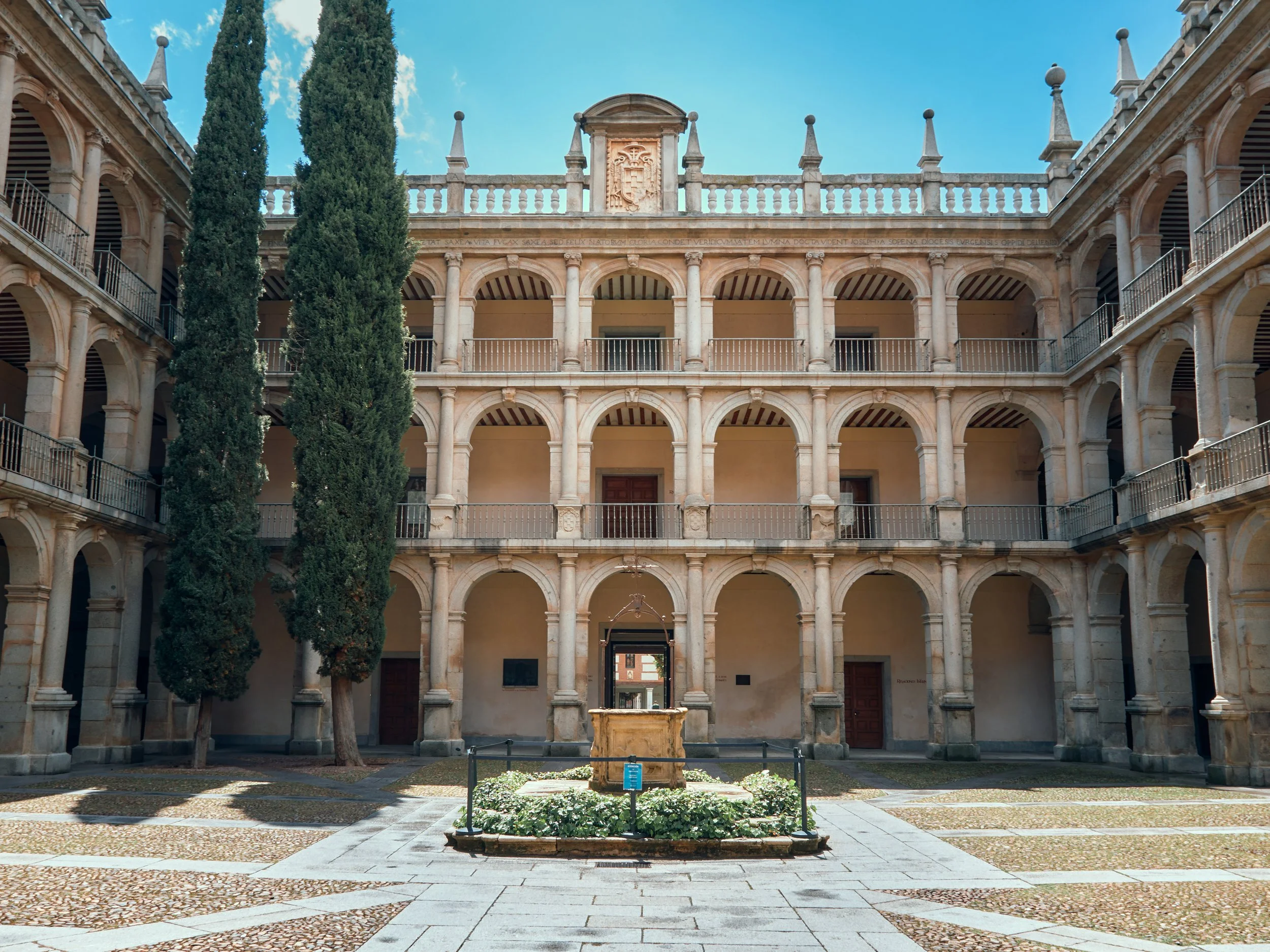 Patio interior de un edificio antiguo de varios pisos con arcos de piedra y un fuente en el centro, flanqueado por dos árboles grandes.