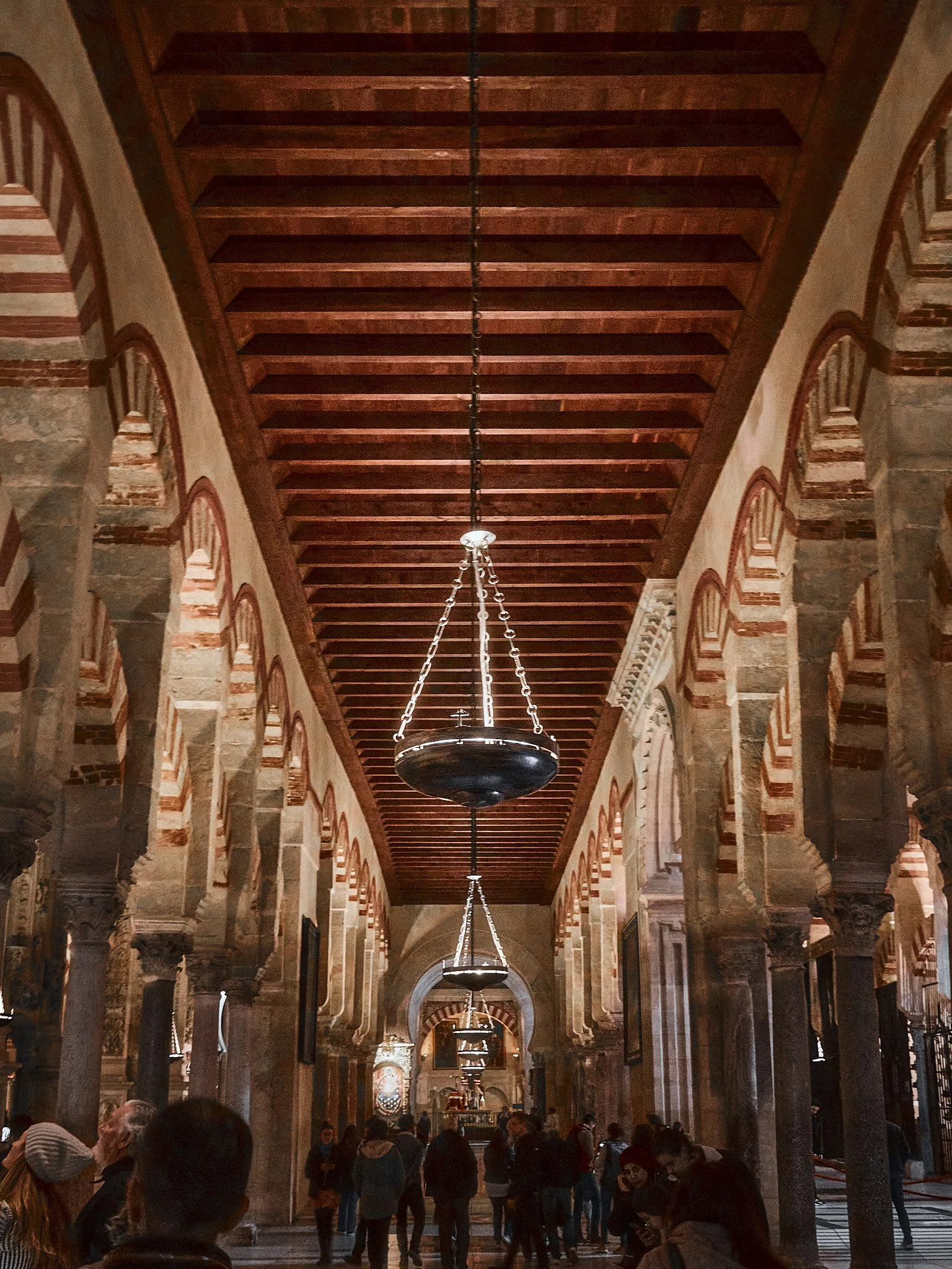 Interior de una iglesia o catedral con columnas de piedra, arcos en las paredes y una serie de lámparas colgantes en el techo de madera. Hay varias personas caminando y en el fondo se observa un altar.