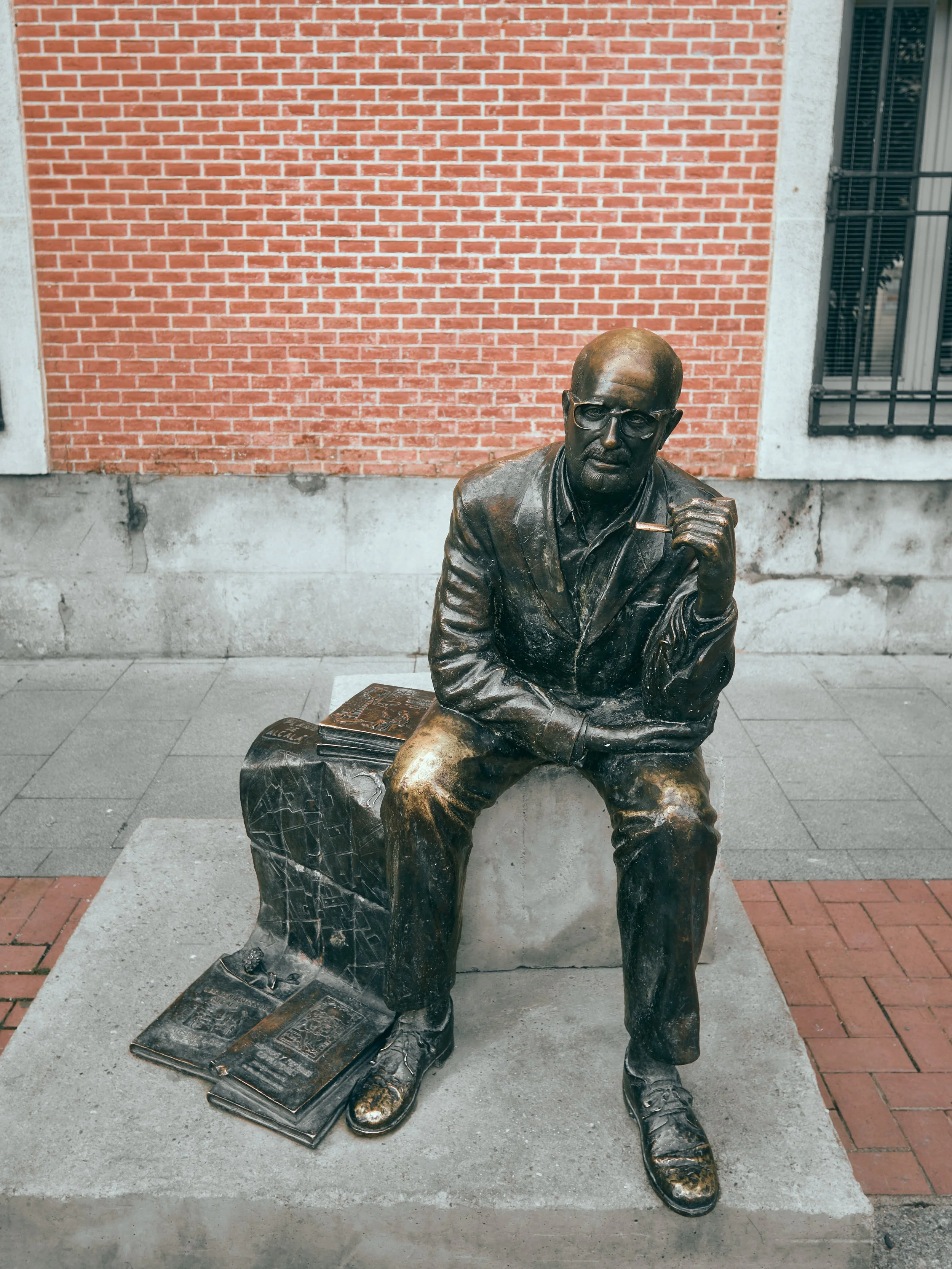 Escultura de un hombre sentado con gafas, con una mano en la mejilla y un cigarrillo en la otra. Tiene libros en el lado izquierdo y otro en el suelo. Fondo de muro de ladrillos y ventanas con rejas.