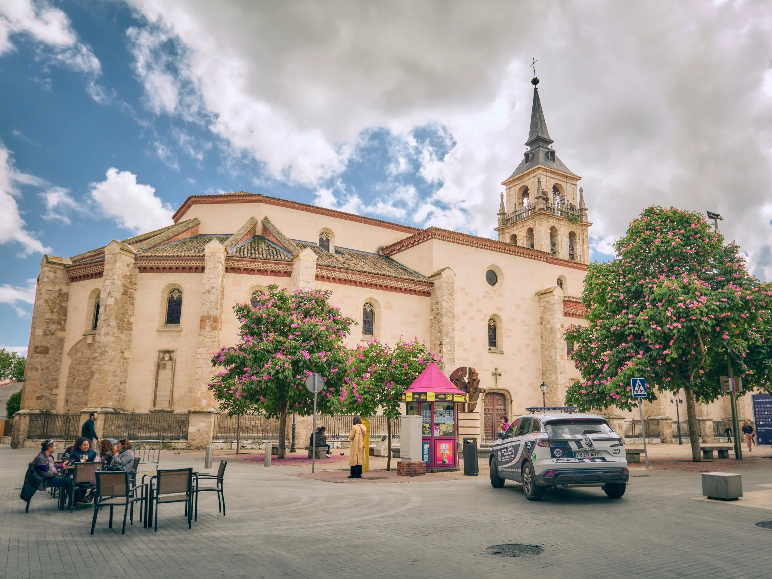 Una iglesia antigua de piedras claras con torres y ventanas góticas en un día soleado con cielo parcialmente nublado, rodeada de árboles con flores rosadas, una plaza con gente sentada en mesas y un coche de policía estacionado frente a la iglesia.