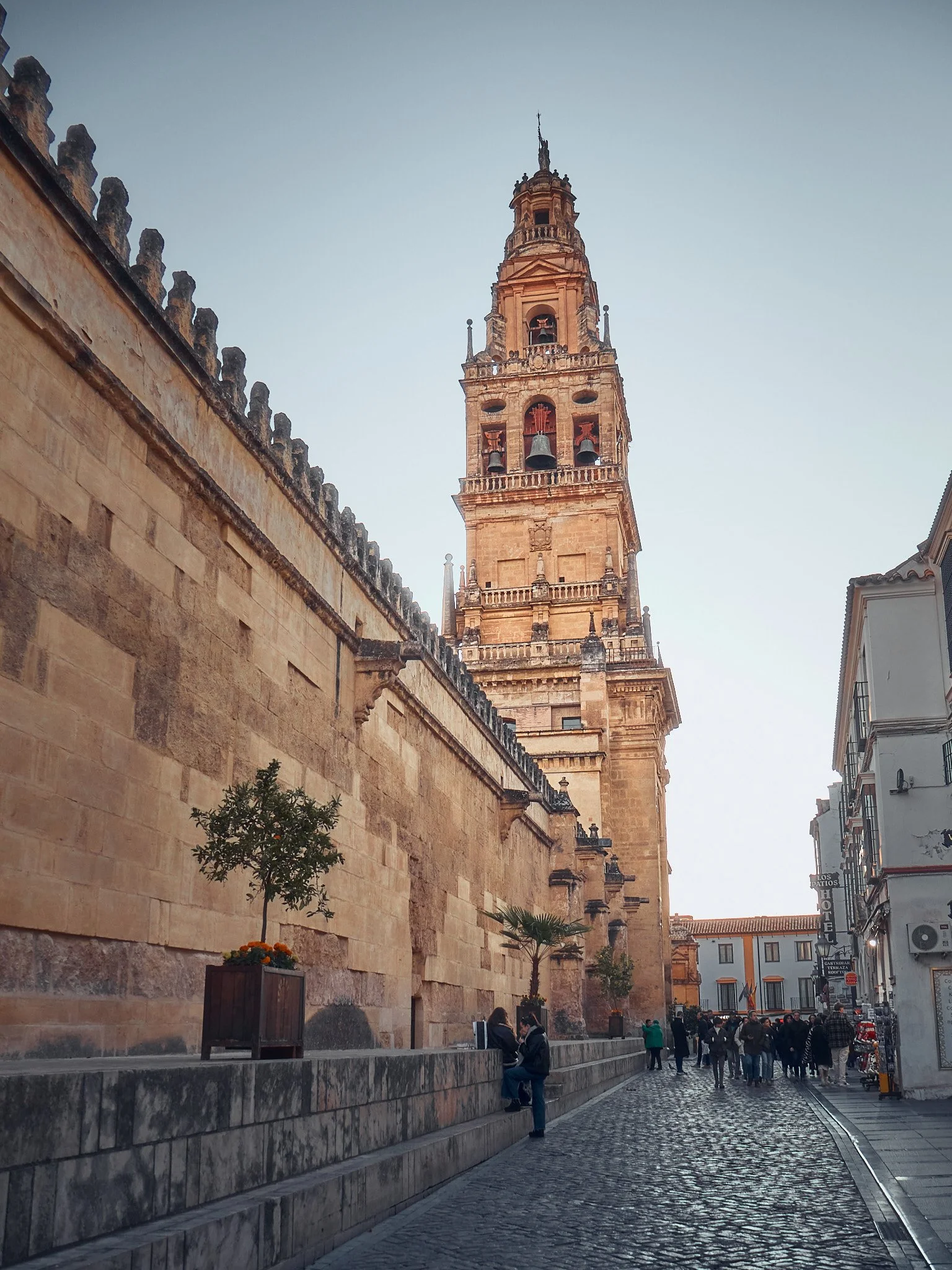 Torre de una catedral antigua, con detalles arquitectónicos en estilo renacentista, en una calle peatonal pavimentada con piedras, con algunas personas sentadas y otras caminando.