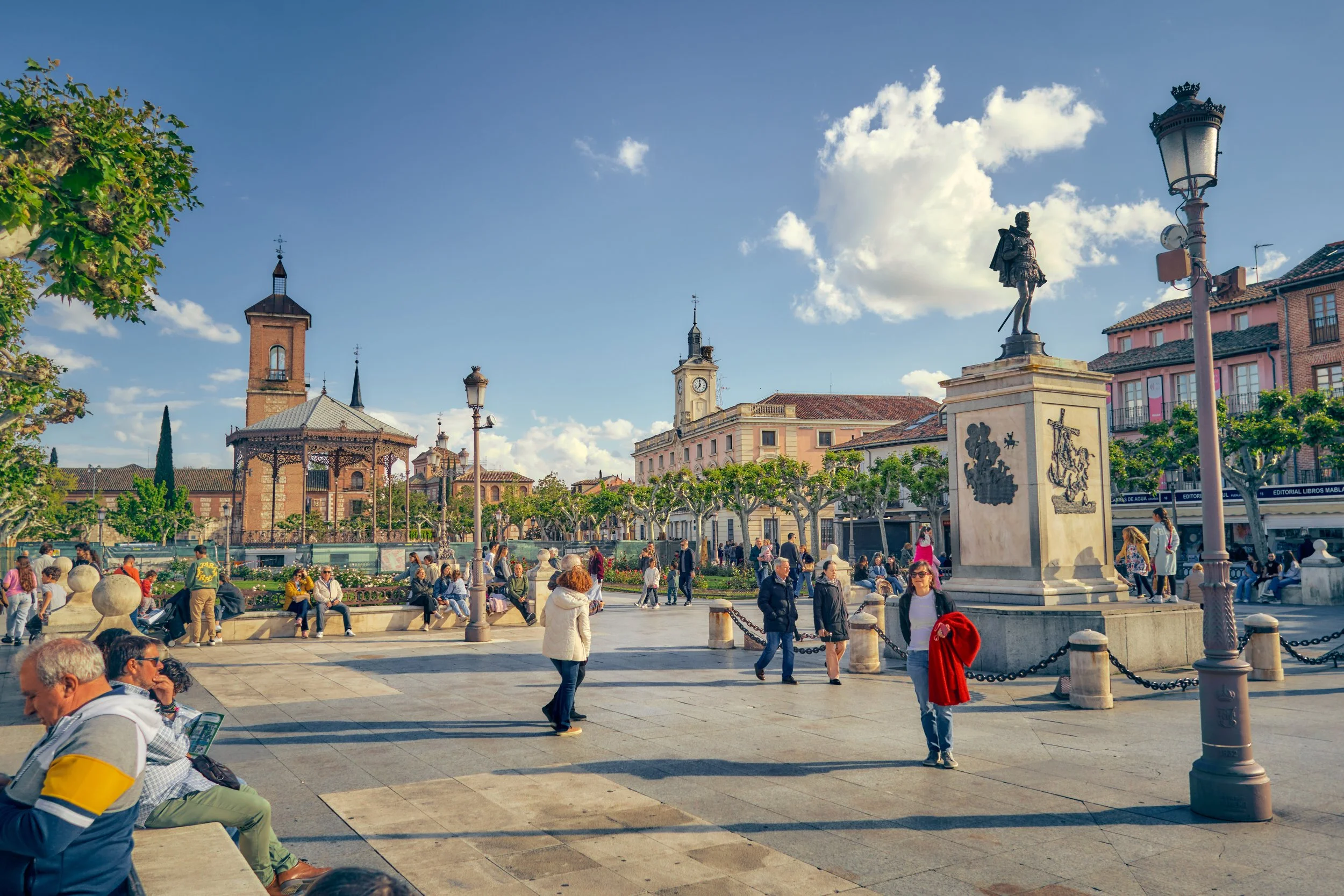 Plaza pública con personas, fuente, monumento y edificios históricos, cielo despejado.
