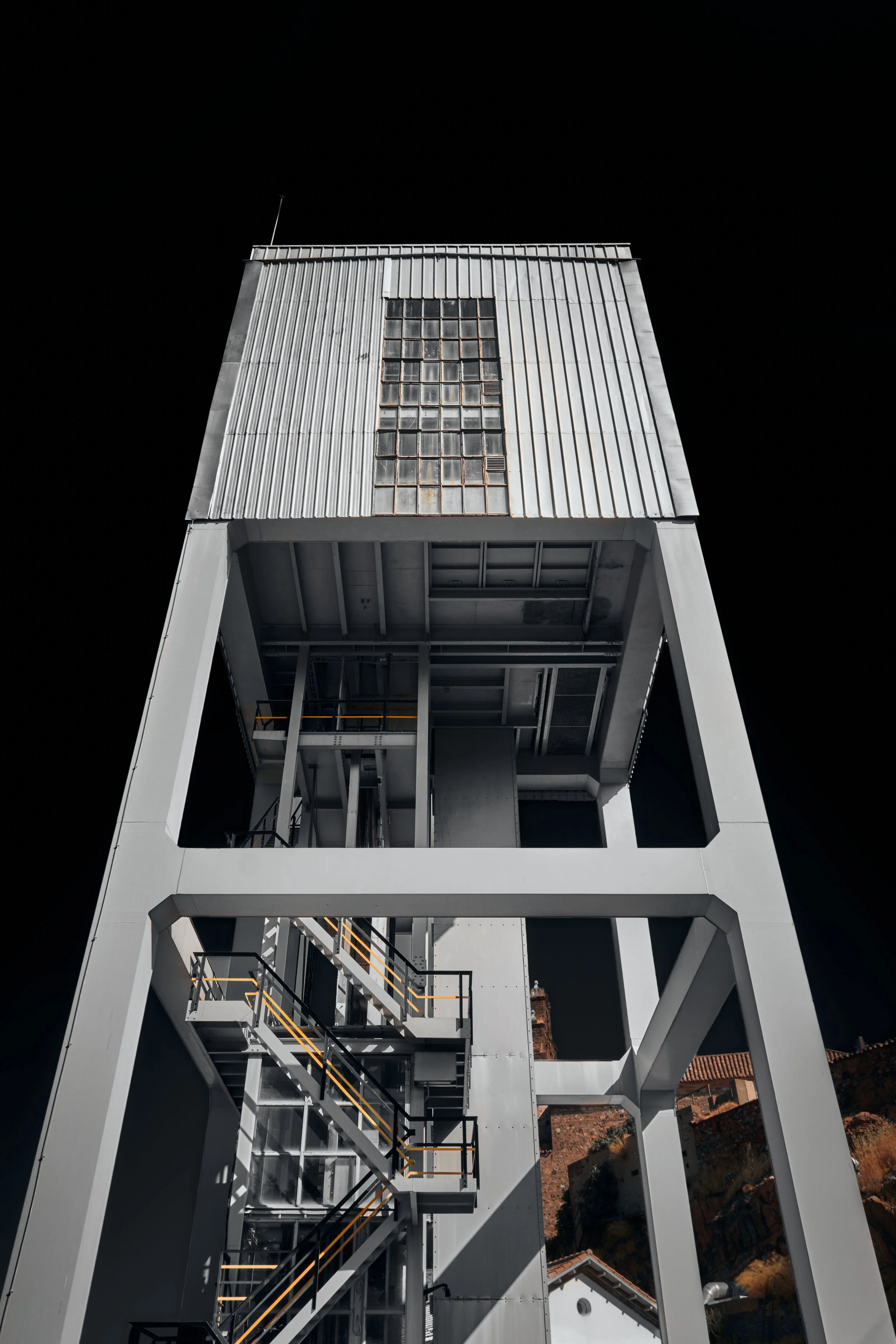 Estructura metálica alta con escaleras y plataforma, con una pared de ventanas en la parte superior, contra un cielo negro, en un entorno urbano o industrial.
