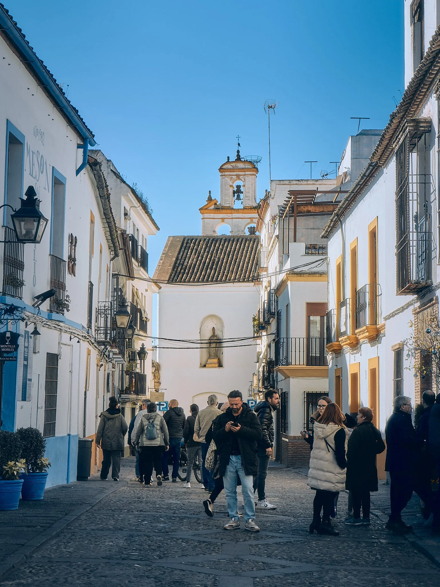 Vista de una calle peatonal en un barrio colonial con personas caminando y edificios blancos con balcones y ventanas con rejas, y una iglesia con campanario al fondo bajo un cielo azul claro.