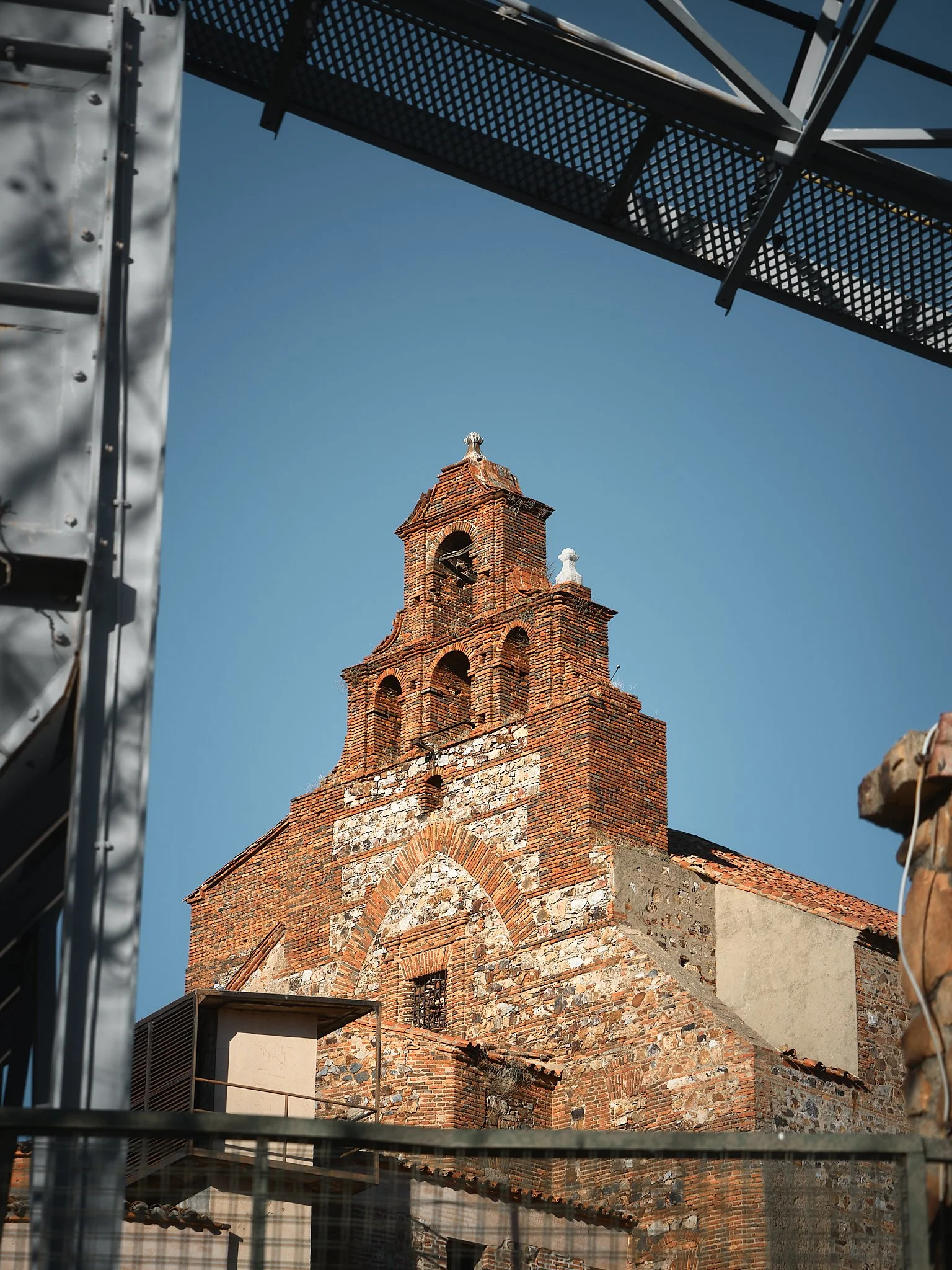 Vista de una iglesia antigua de ladrillo y piedra, enmarcada por estructuras metálicas en primer plano, con un cielo despejado de fondo.