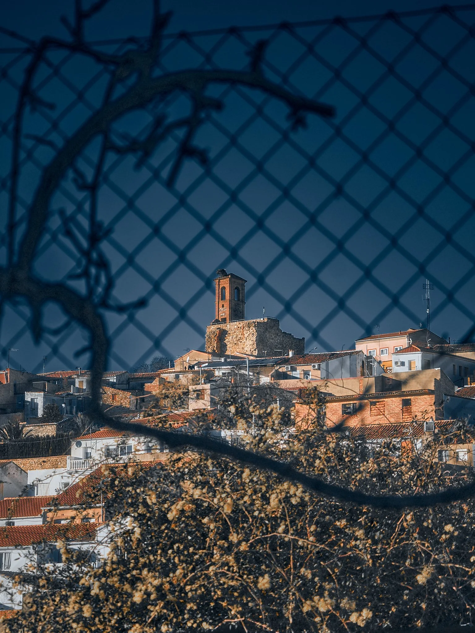 Vista de un pueblo con casas y techos rojos, y una torre antigua en el fondo, vista a través de una reja de metal.