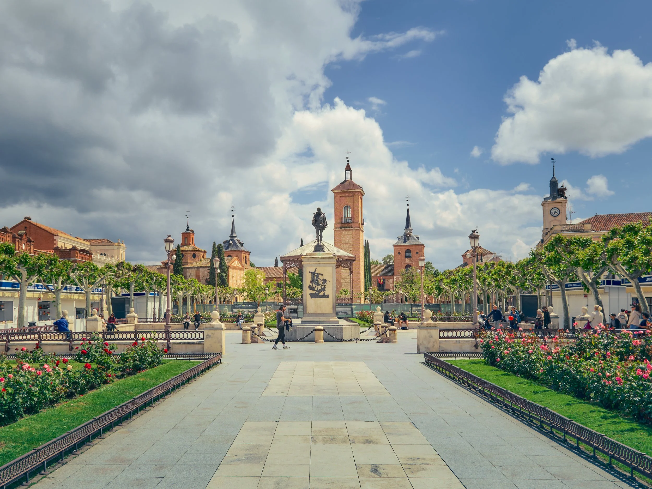Plaza con fuente y monumento en el centro, rodeada de árboles podados y bancos, con personas caminando y sentadas, edificios históricos y reloj en la esquina, bajo cielo parcialmente nublado.