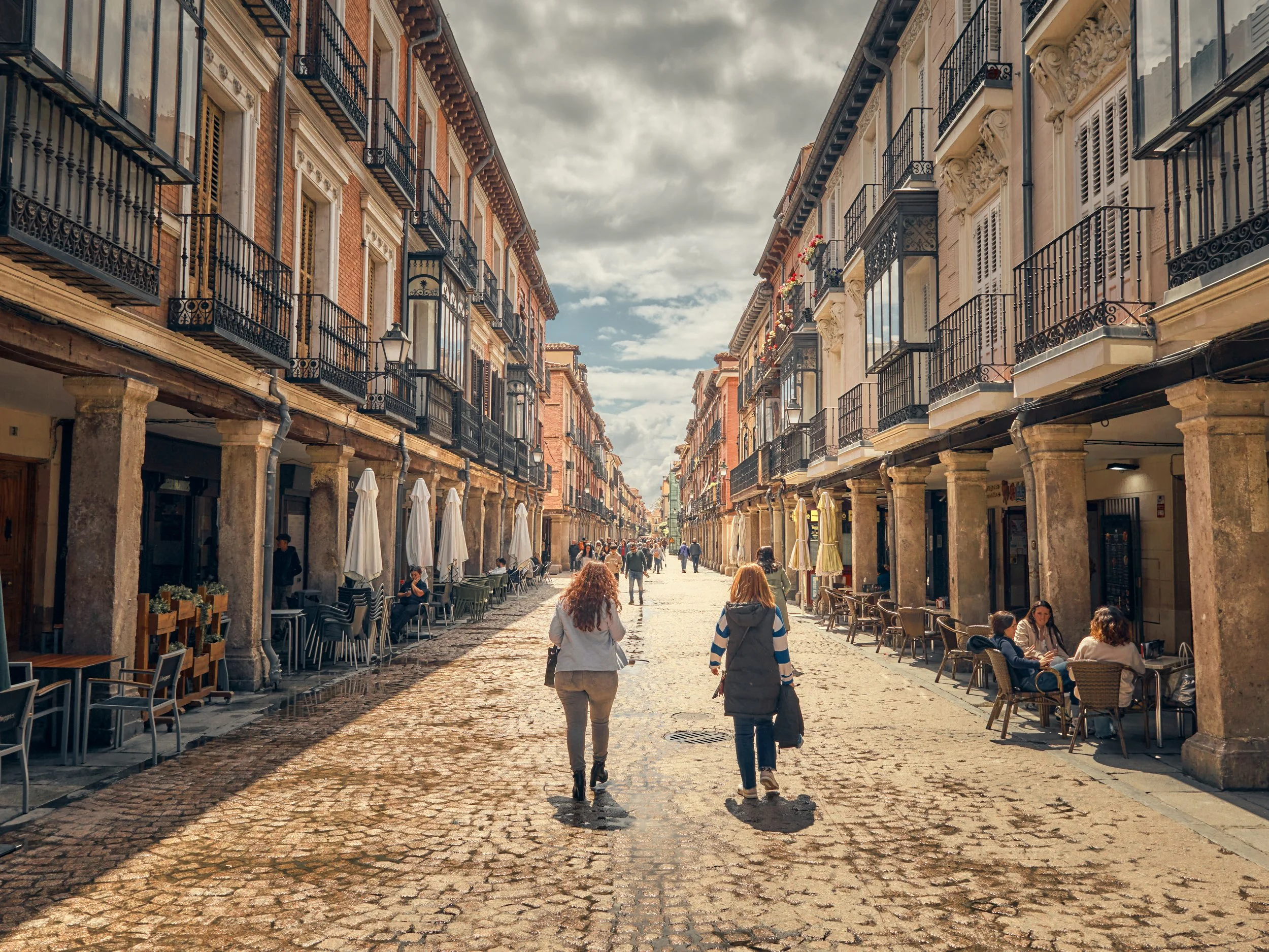 Avenida peatonal con edificios históricos de fachadas coloniales, algunas con balcones de hierro forjado, en una ciudad de arquitectura europea. Hay personas caminando y sentadas en terrazas de cafés y restaurantes, bajo un cielo parcialmente nublado