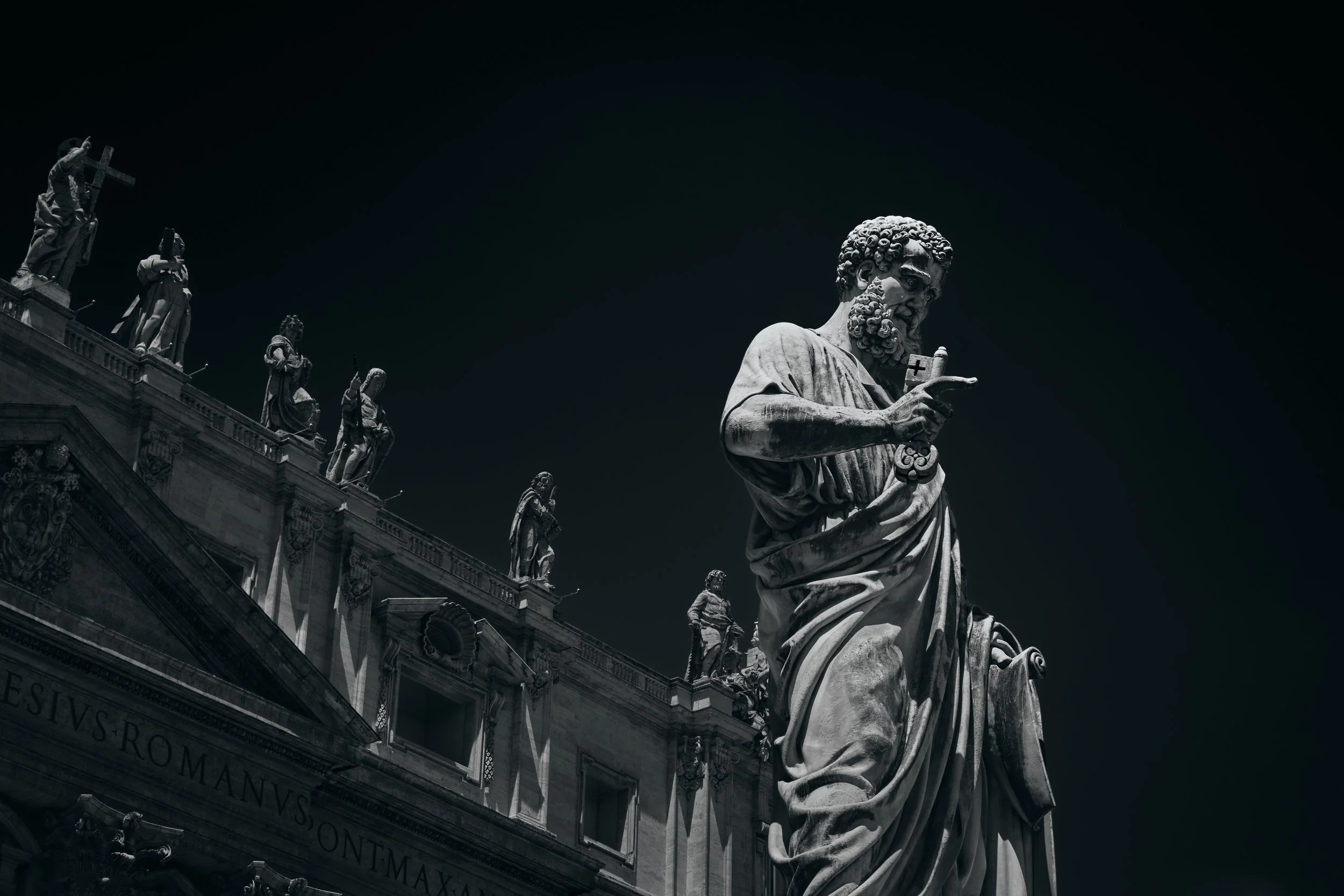 Estatua clásica en primer plano con varias estatuas en la azotea y fondo oscuro