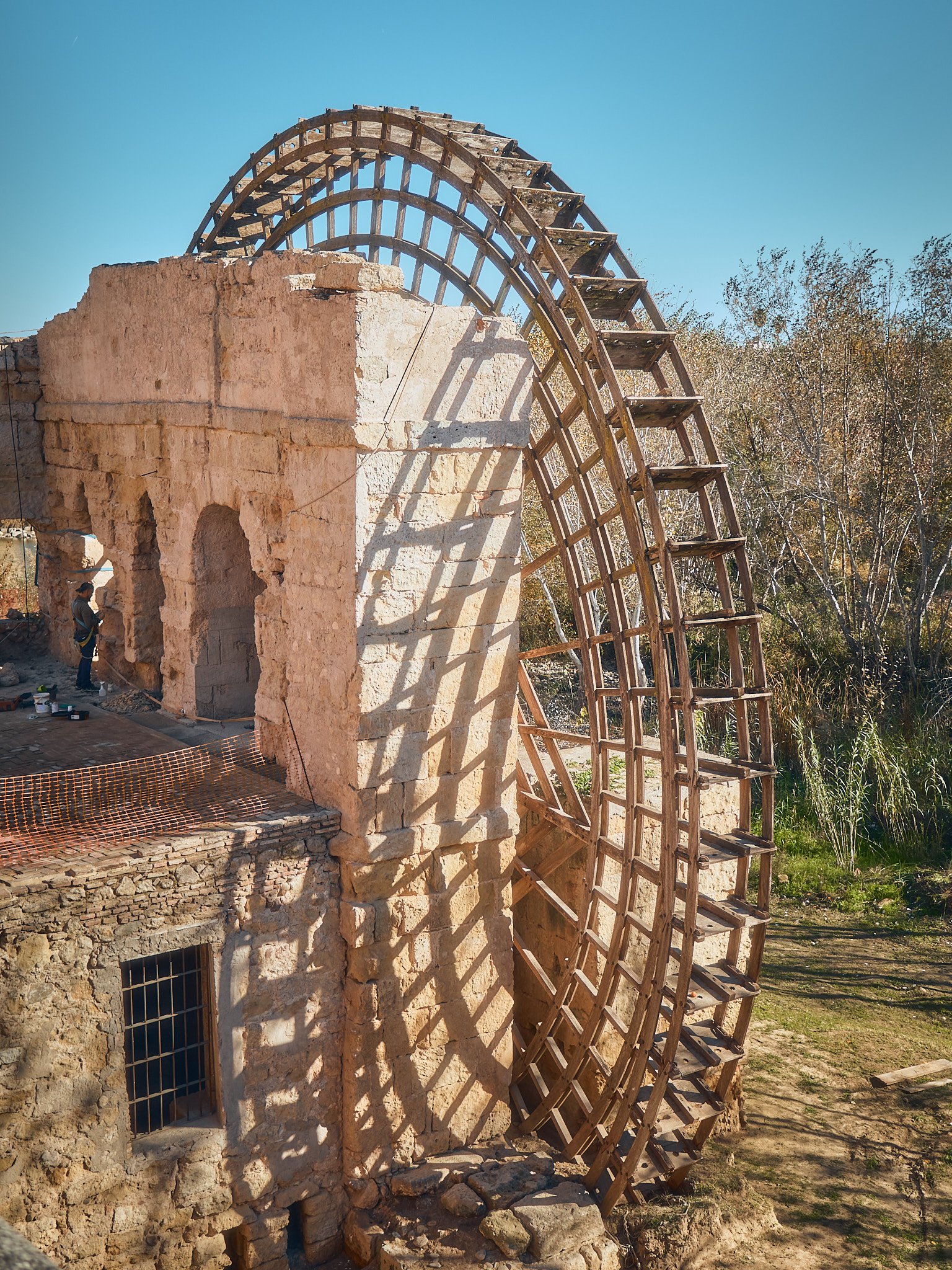 Rueda de agua en construcción en una estructura de piedra con sombra en el suelo y árboles en el fondo.