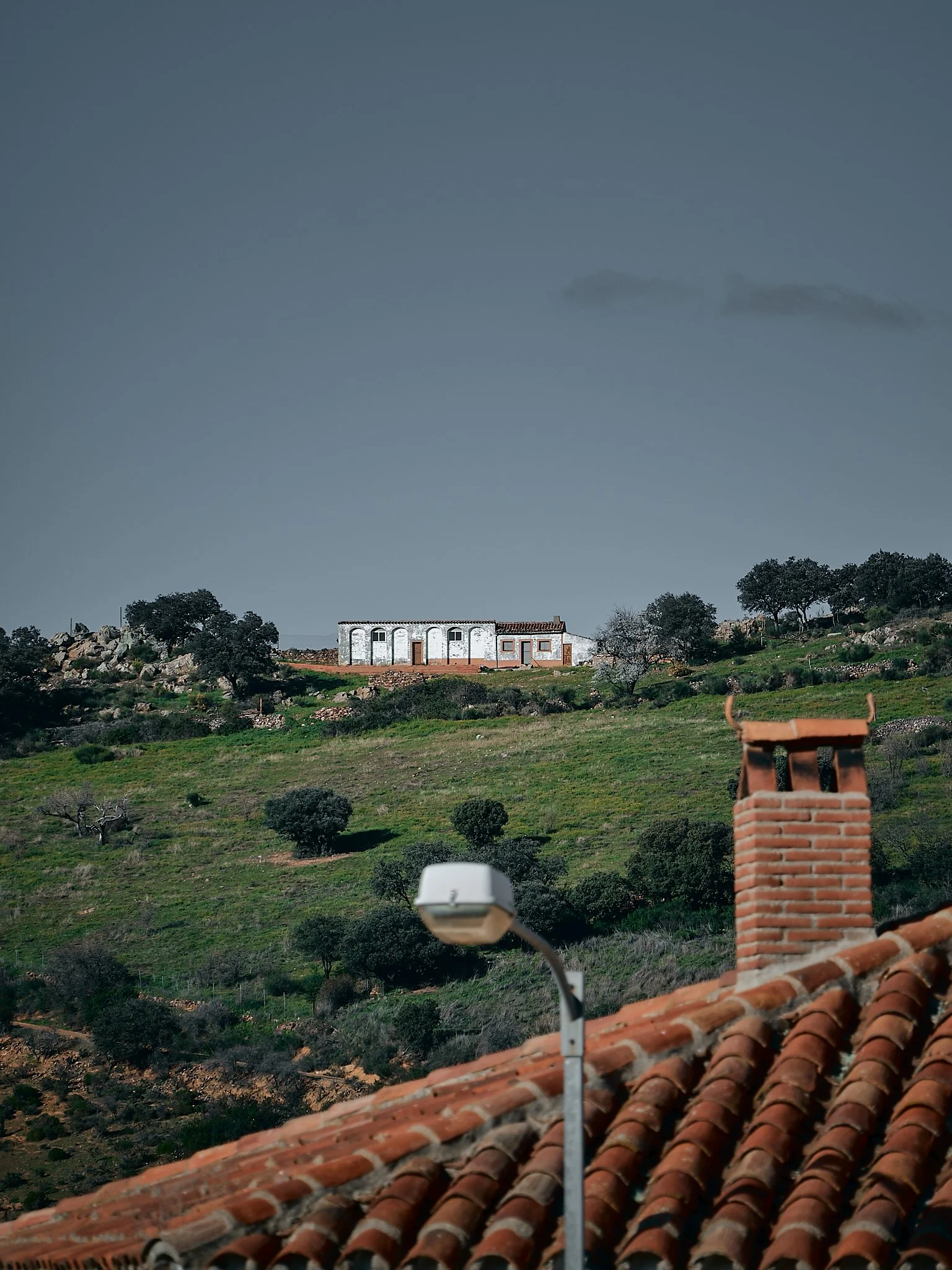 Vista de un paisaje rural con casas blancas en una colina, árboles dispersos, colinas verdes, un poste de luz y un techo de tejas en primer plano.