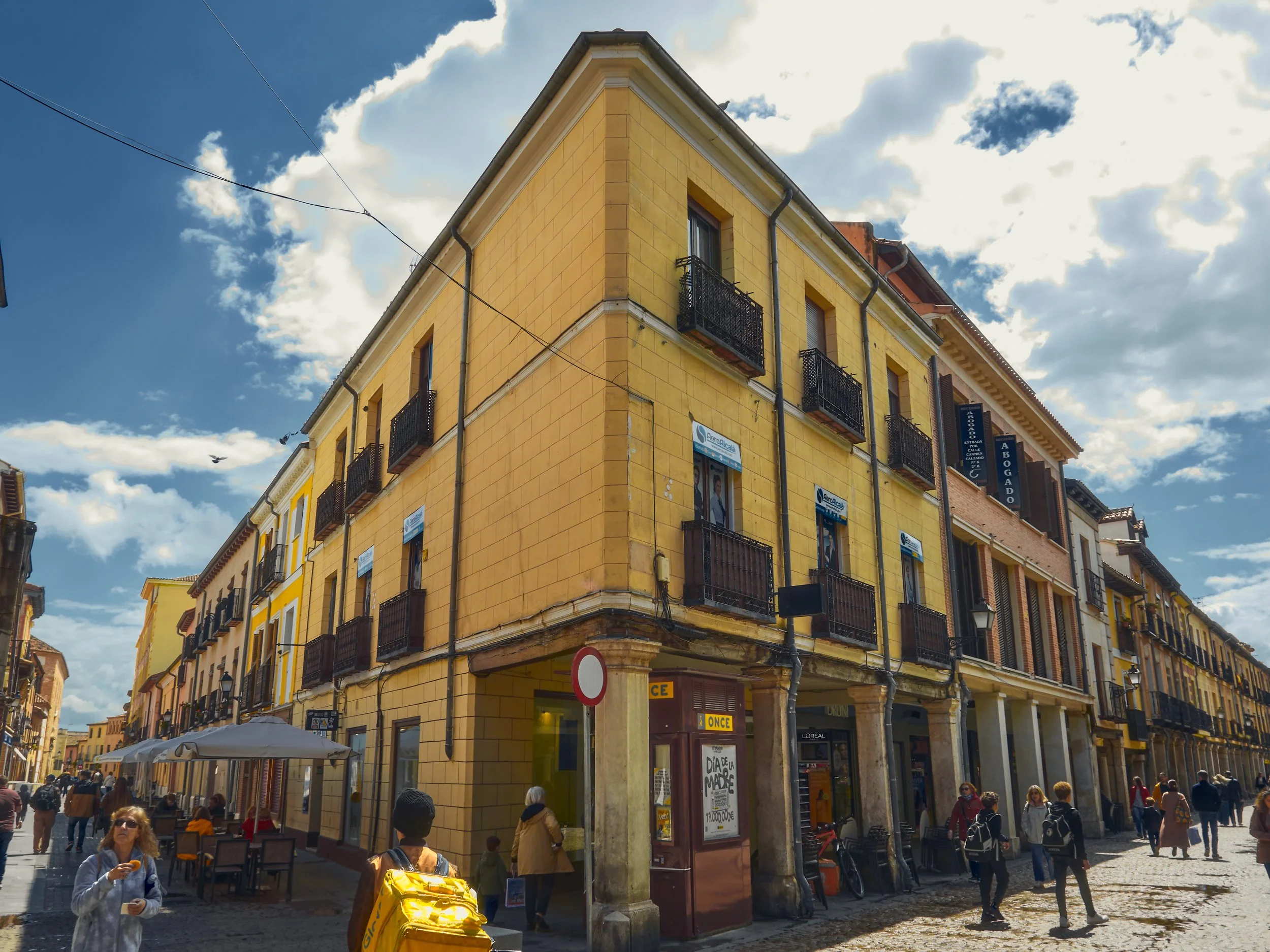 Edificio de color amarillo en una calle peatonal, con varias personas caminando y sentadas en mesas de café, con un cielo parcialmente nublado.